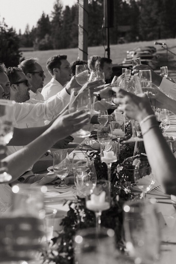 People raising glasses in a toast at a long outdoor reception table during daytime.