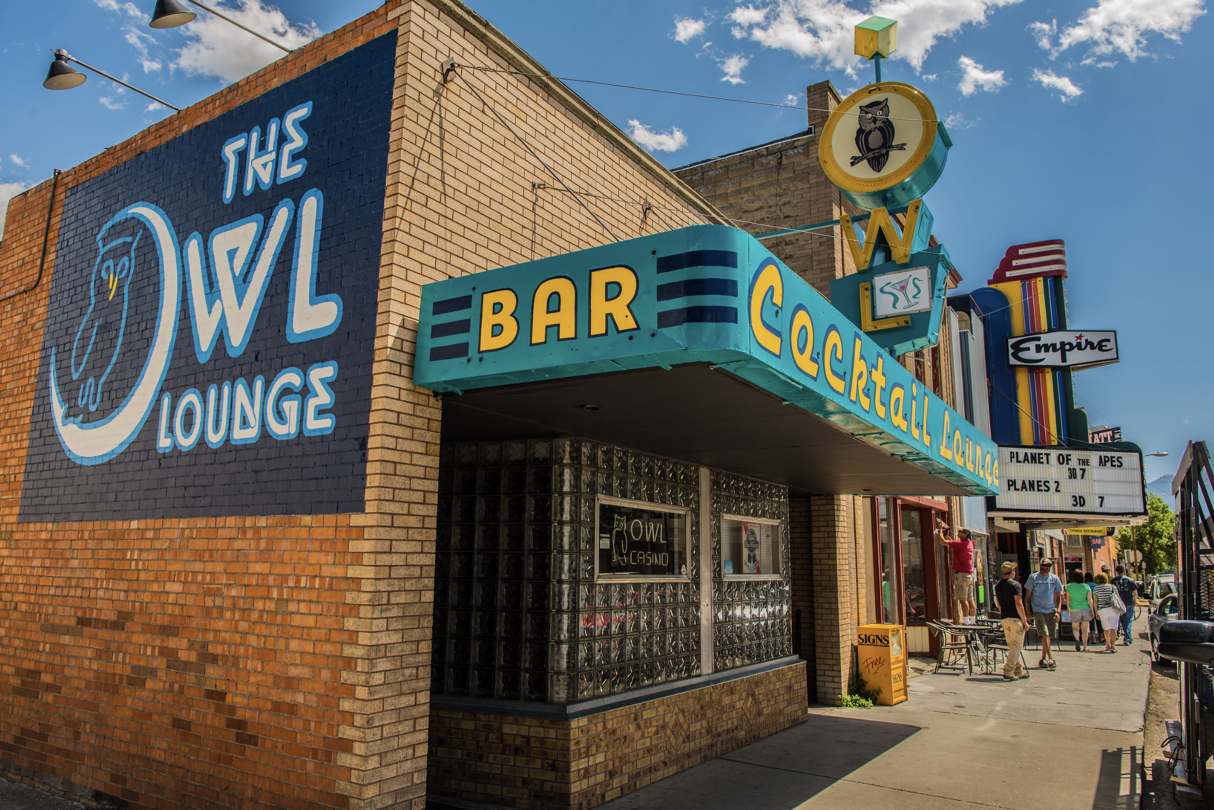 Street view of The Owl Lounge, a bar and cocktail lounge with neon signs, including a large owl logo, a marquee sign, and people walking on the sidewalk on a sunny day.