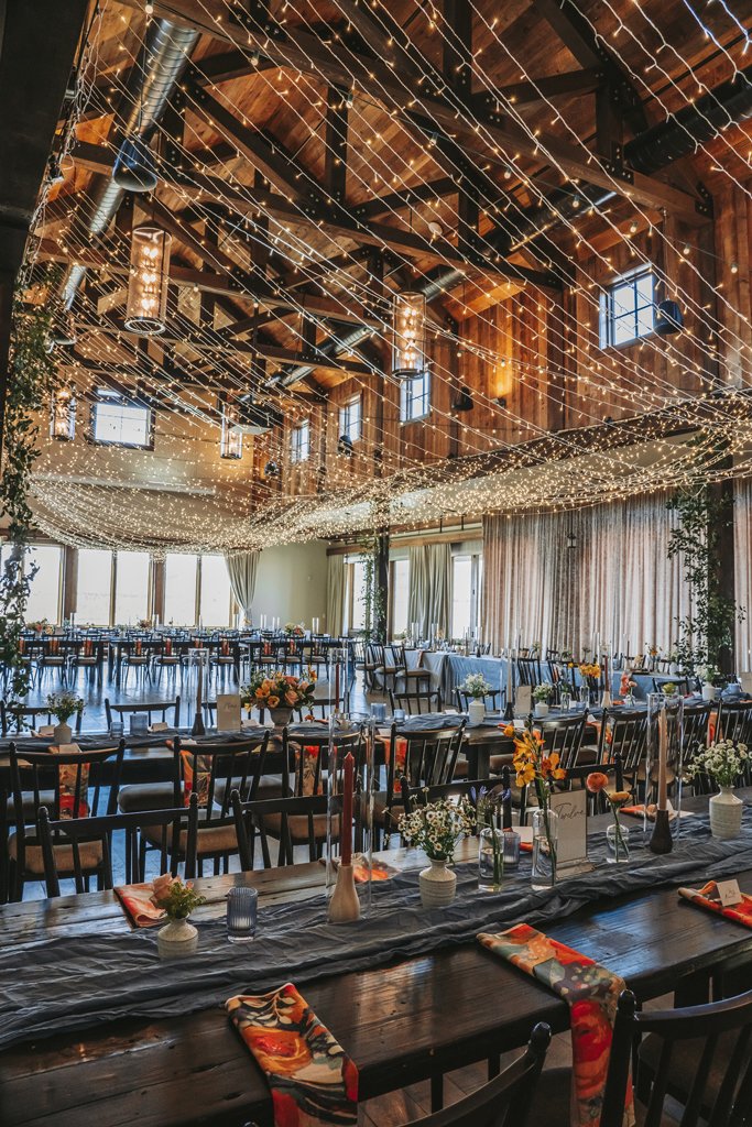 Decorated banquet hall with string lights hanging from wooden ceiling, featuring long tables with flower centerpieces, candles, and place settings, large windows with curtains, and a rustic ambiance for a celebration or wedding reception.
