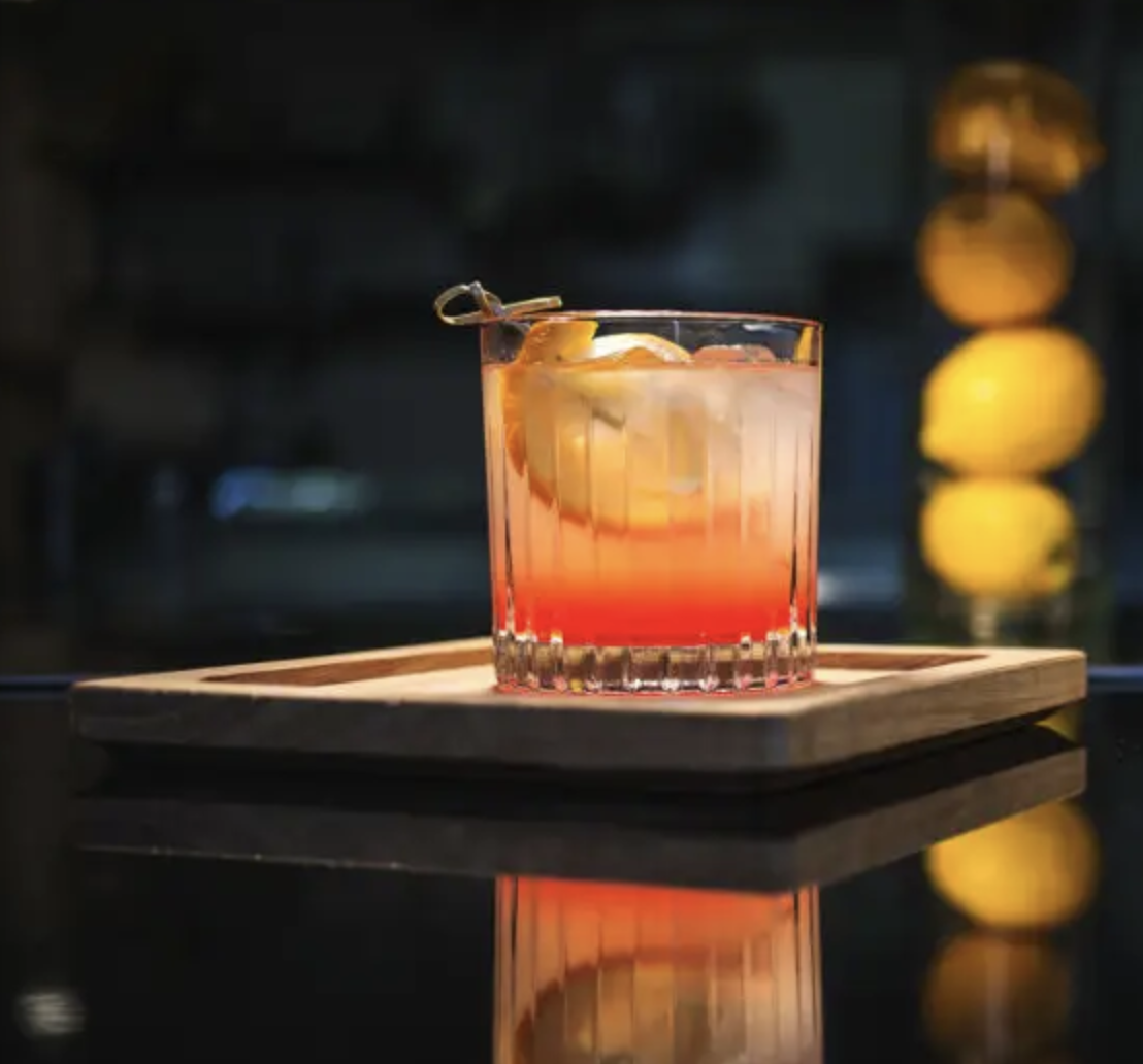A cocktail with a red and orange gradient color, garnished with lemon slices, in a clear glass on a wooden tray, with blurry yellow lights in the background.