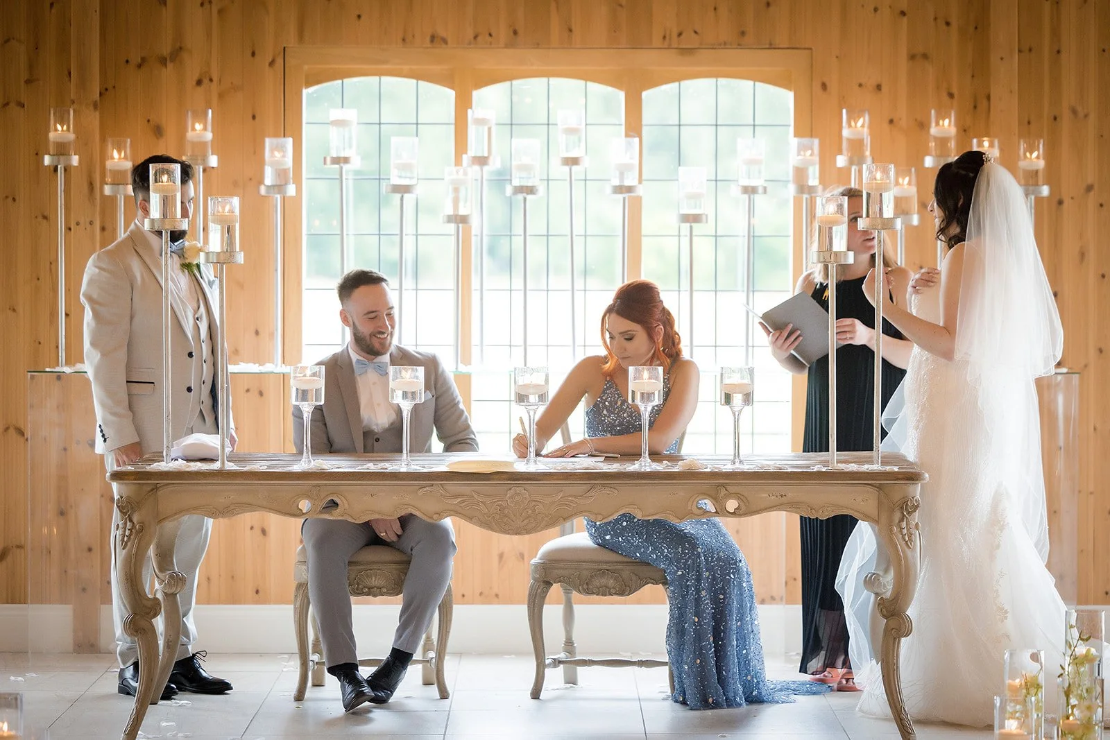 A wedding ceremony with five people inside a wooden-paneled room. The bride and groom are standing and sitting at a table with candles and glassware. The bride is in a white wedding dress and veil, and the groom is in a light suit. Two women are stan