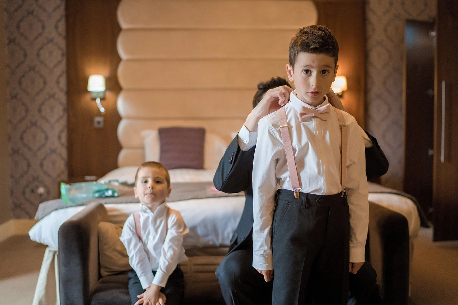 Page boy helping groom adjust bow tie during preparations, The Mere hotel room
