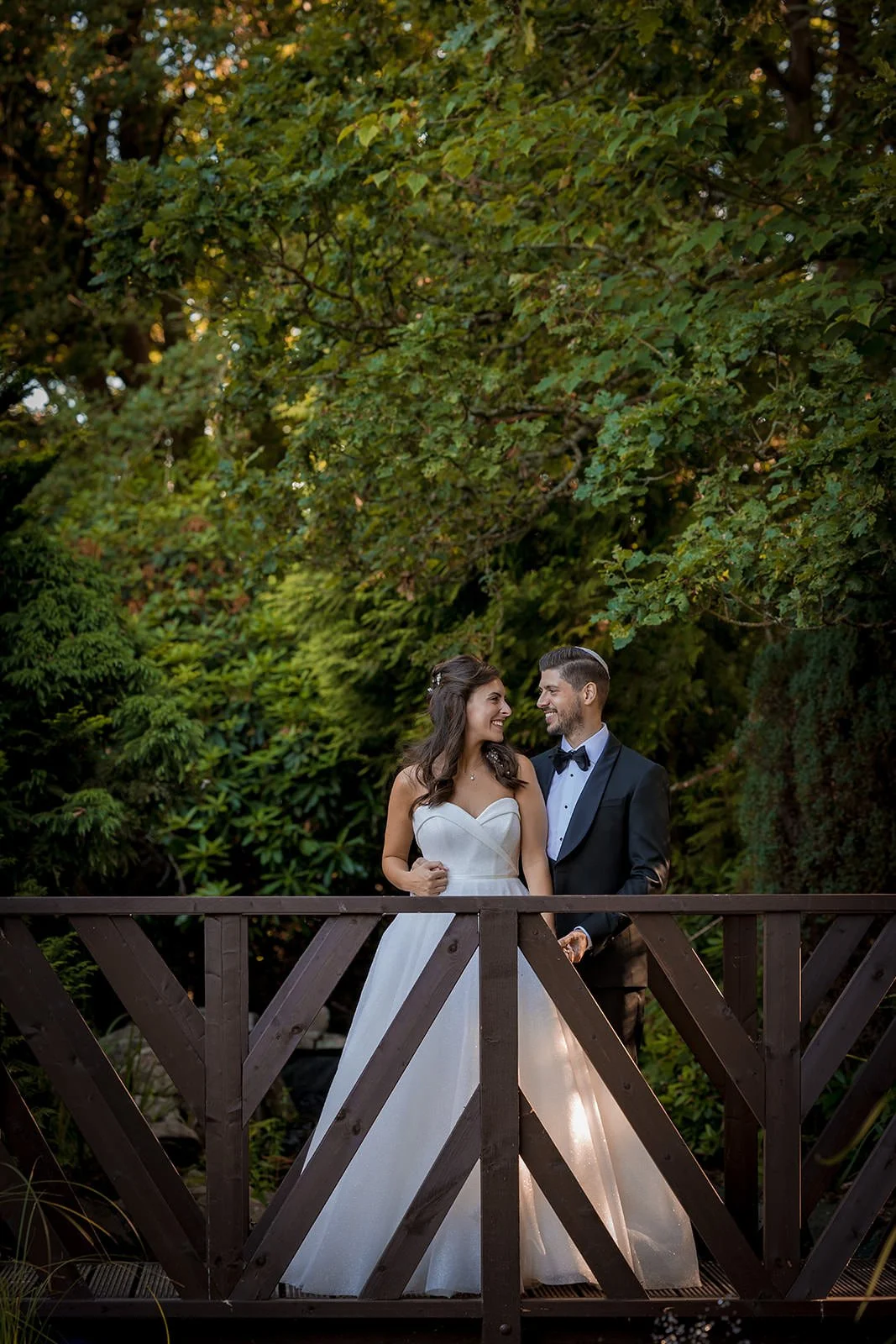 Bride and groom on wooden bridge in Nunsmere Hall gardens, green foliage behind
