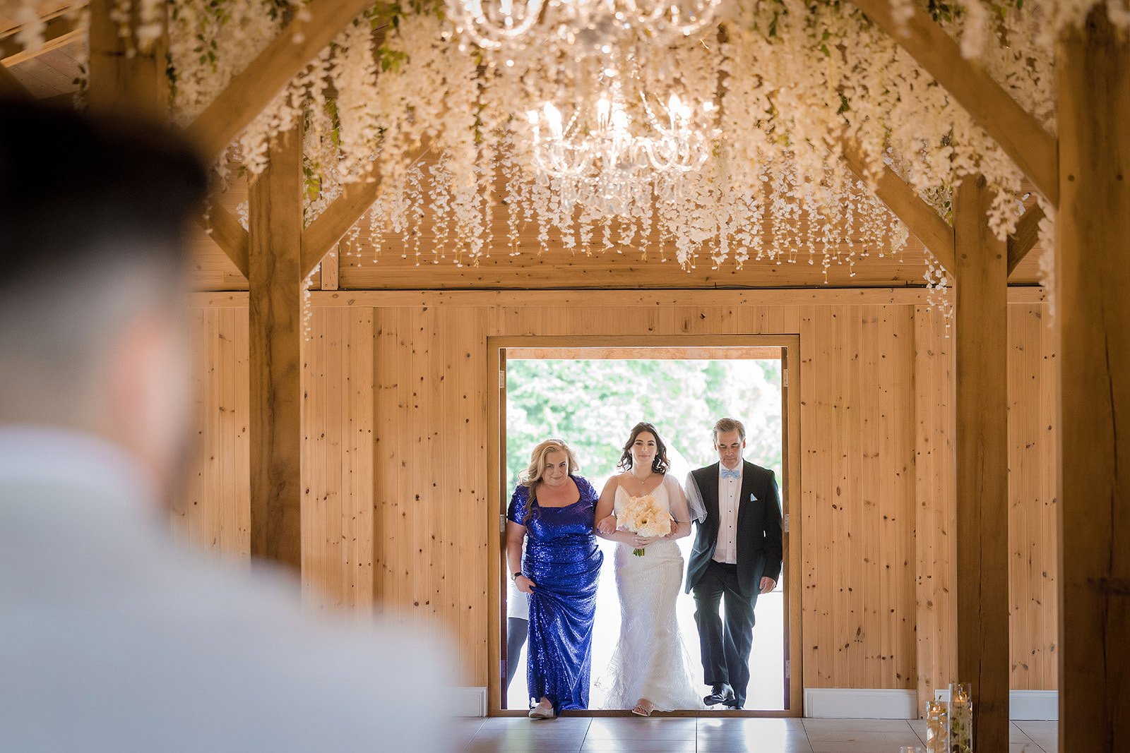 view overlooking groom's shoulder of bride and parents make entrance to the wedding ceremony aisle