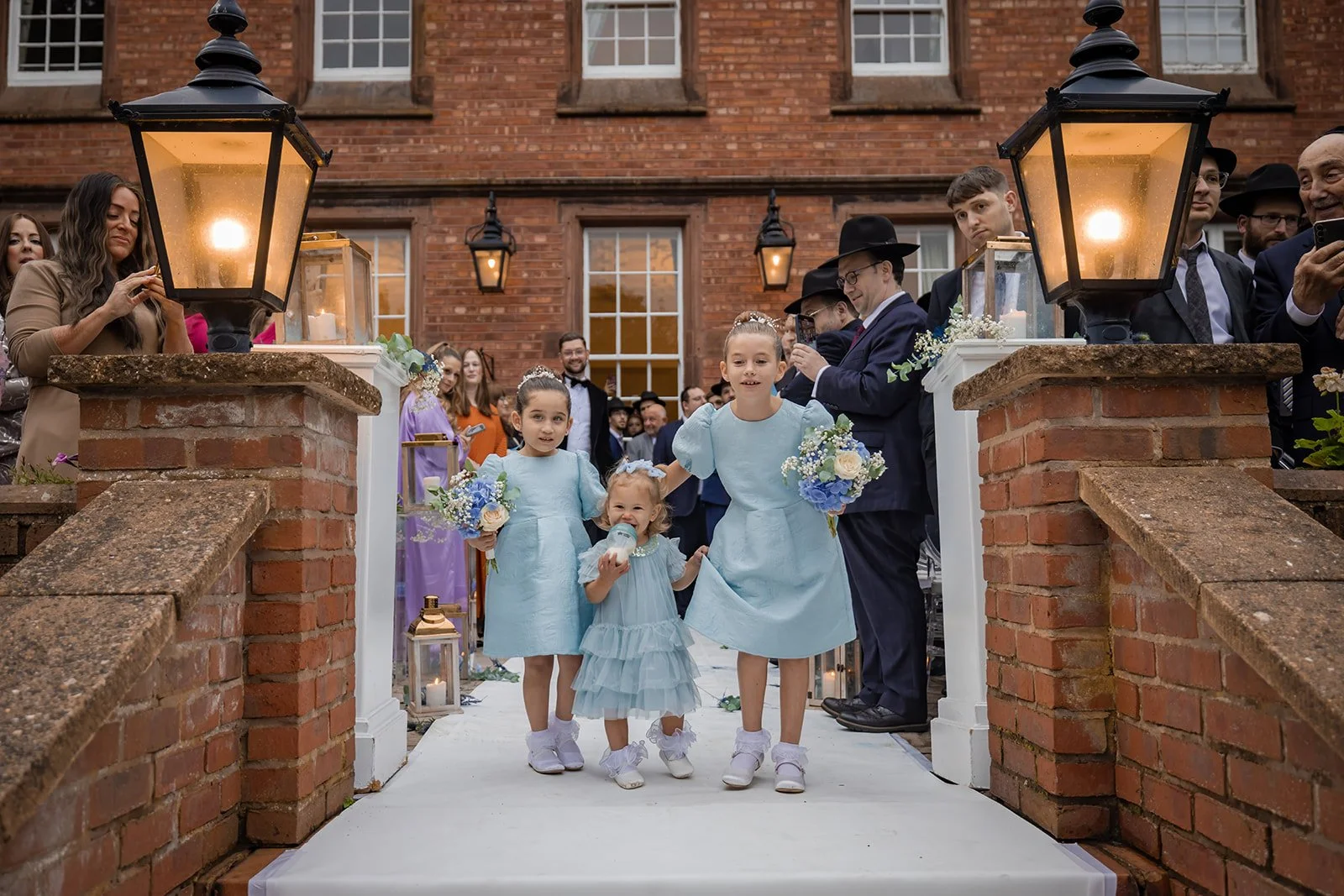 Bride arriving at Nunsmere Hall entrance with flower girls, red brick manor facade
