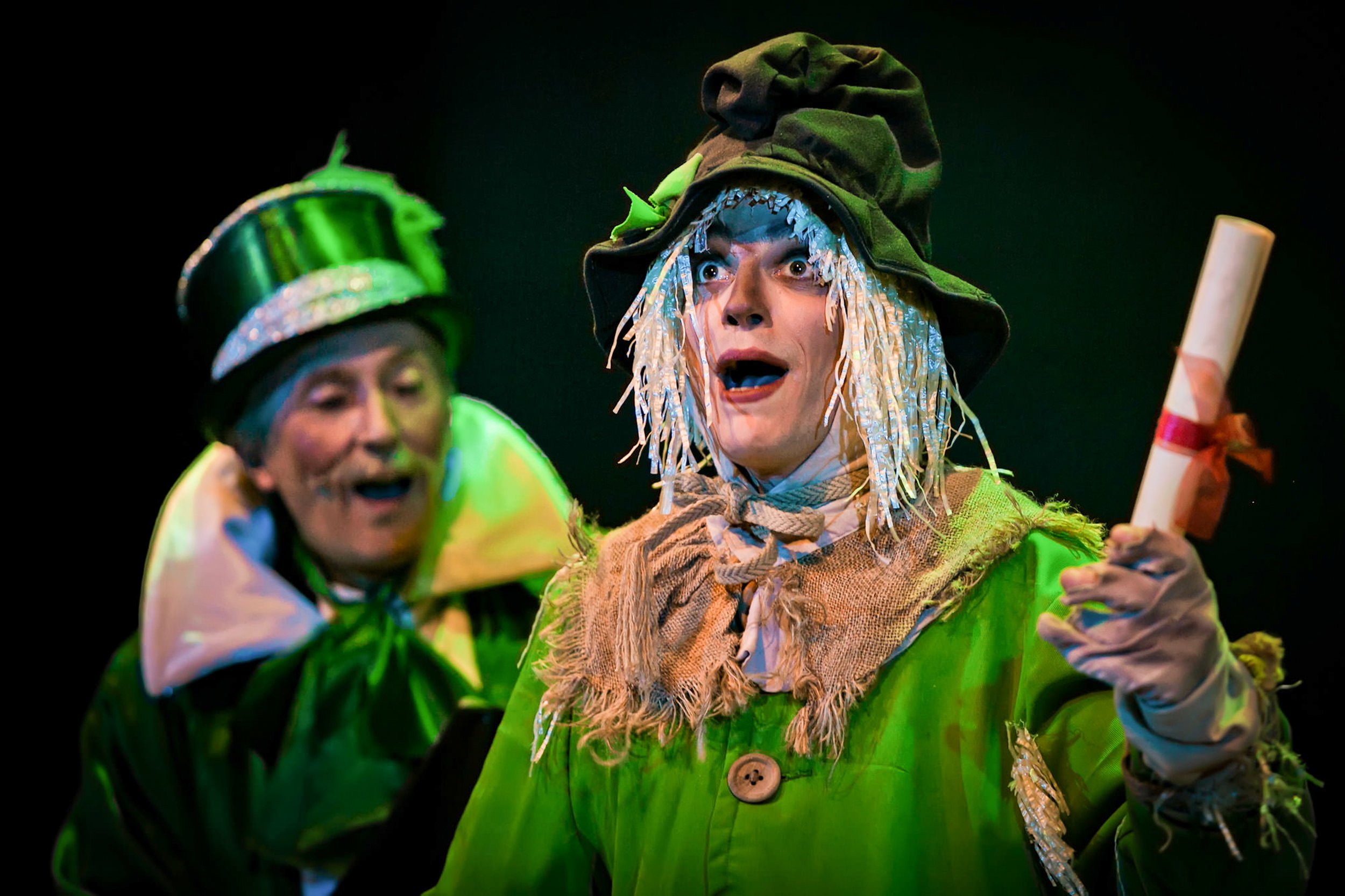 Actors dressed in colorful costumes perform on stage, with one person at the center wearing a green outfit and a hat with long, white wig-like hair, holding a rolled-up paper.