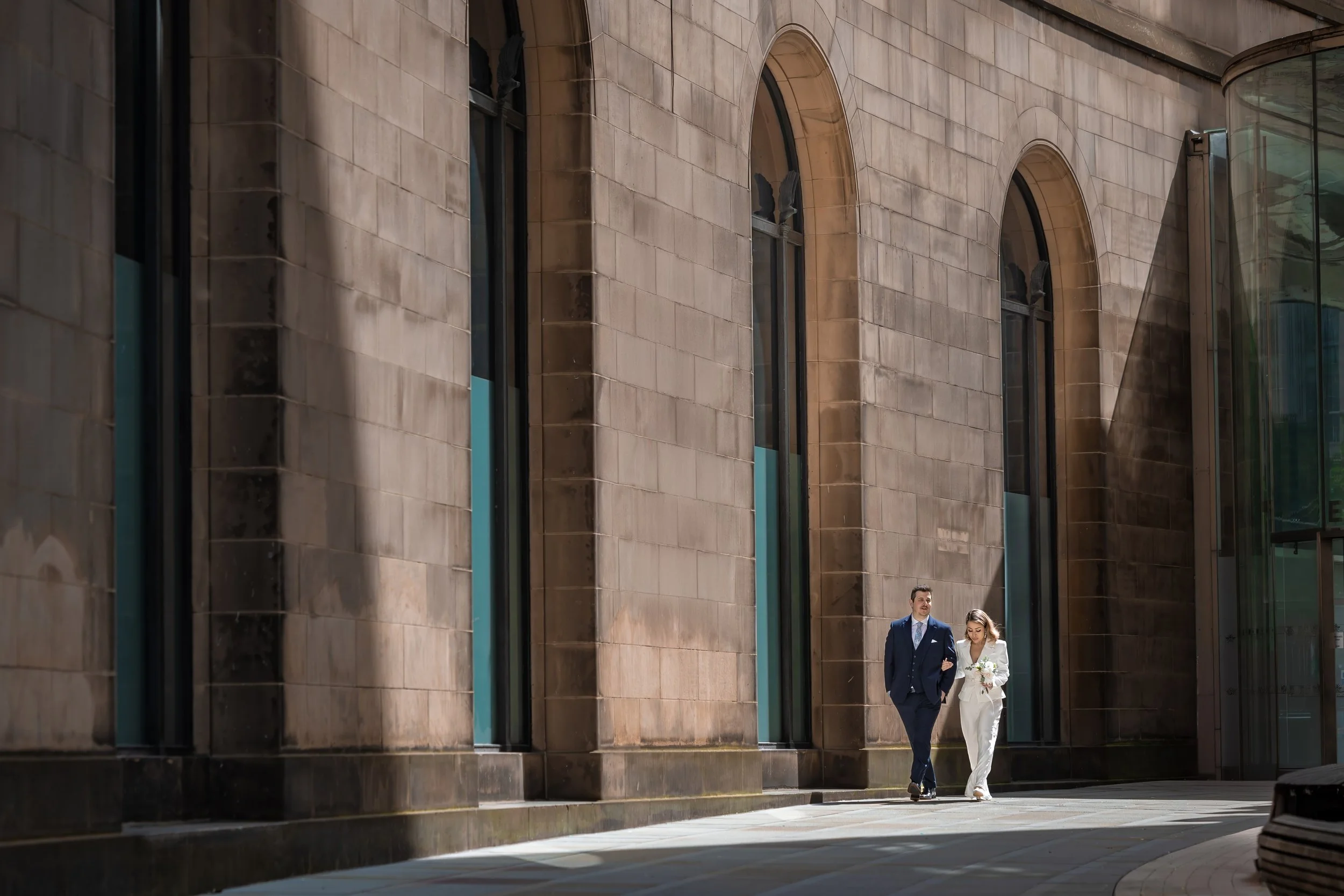 A man in a suit and a woman in a white dress with a bouquet walk together outside a modern building with large arched windows and beige stone walls.