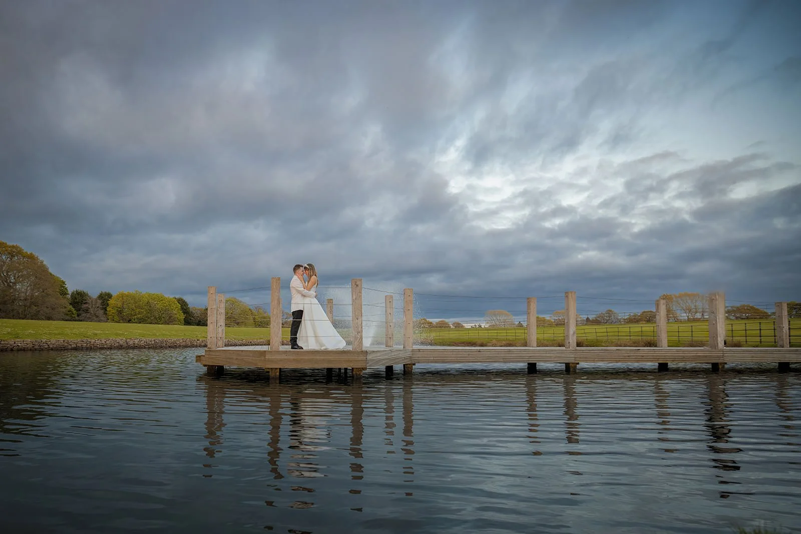 A bride and groom at Merrydale Manor standing on a wooden jetty over water, embracing and kissing, with a cloudy sky and green landscape in the background.