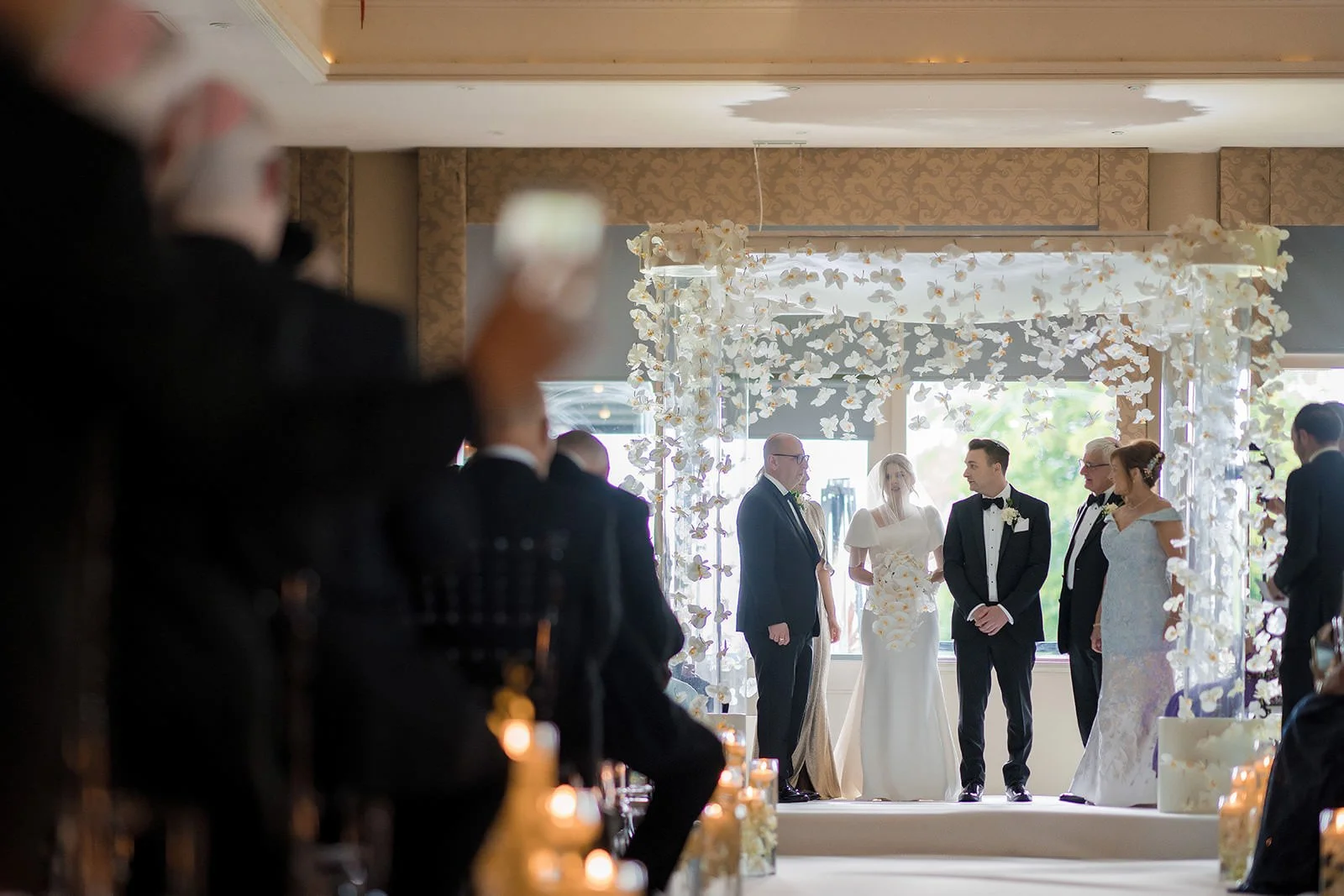 Groom and guests at chuppah with tall windows behind, The Mere ceremony suite

