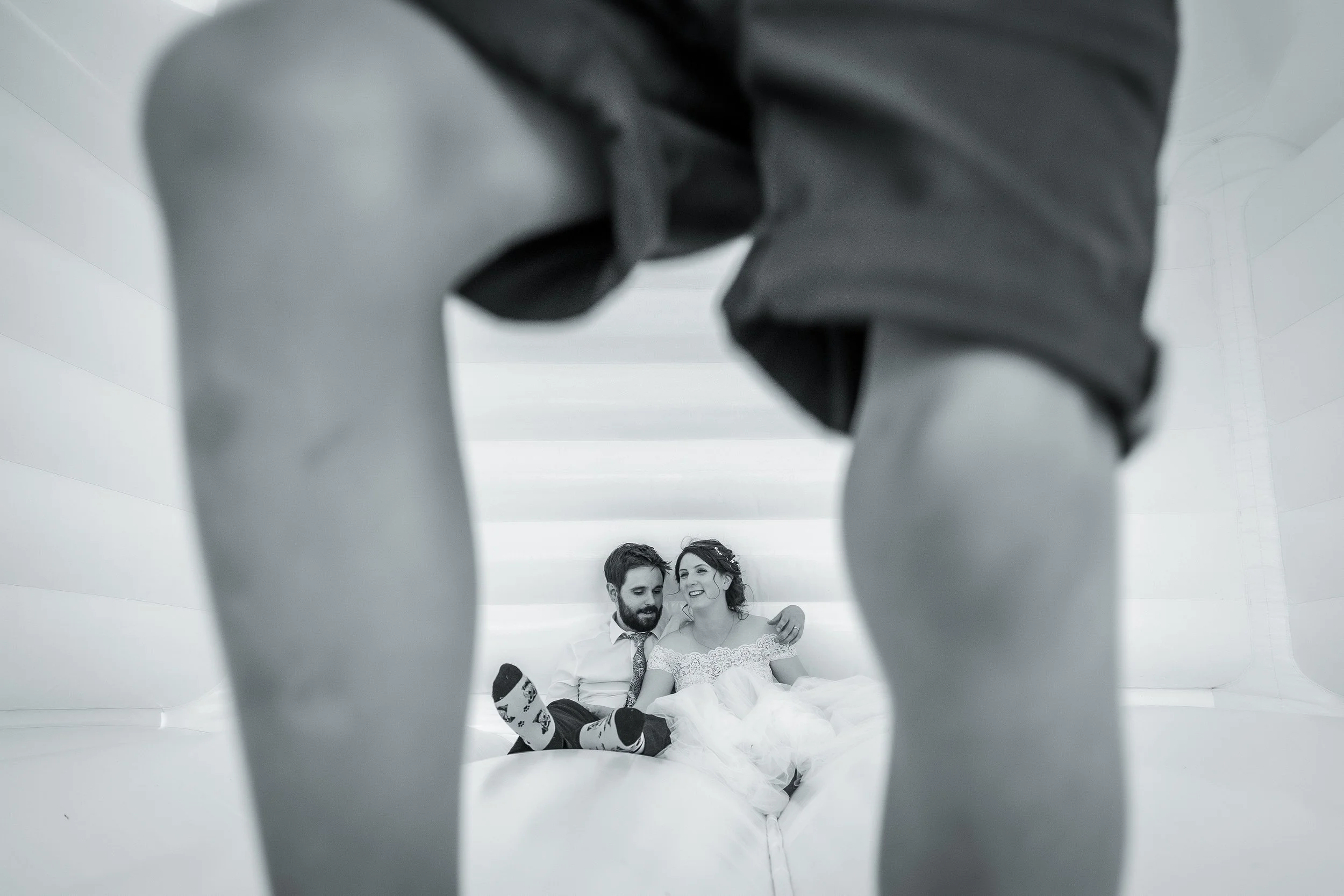 candid photo of bride and groom resting on bouncy castle photographed through child's legs