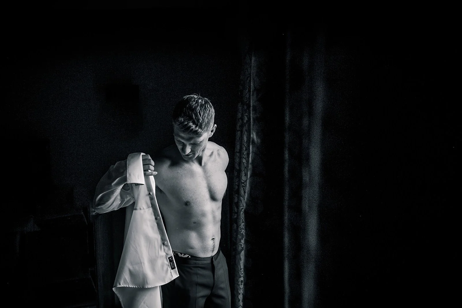 Groom putting his shirt on by window with natural light during groom prep