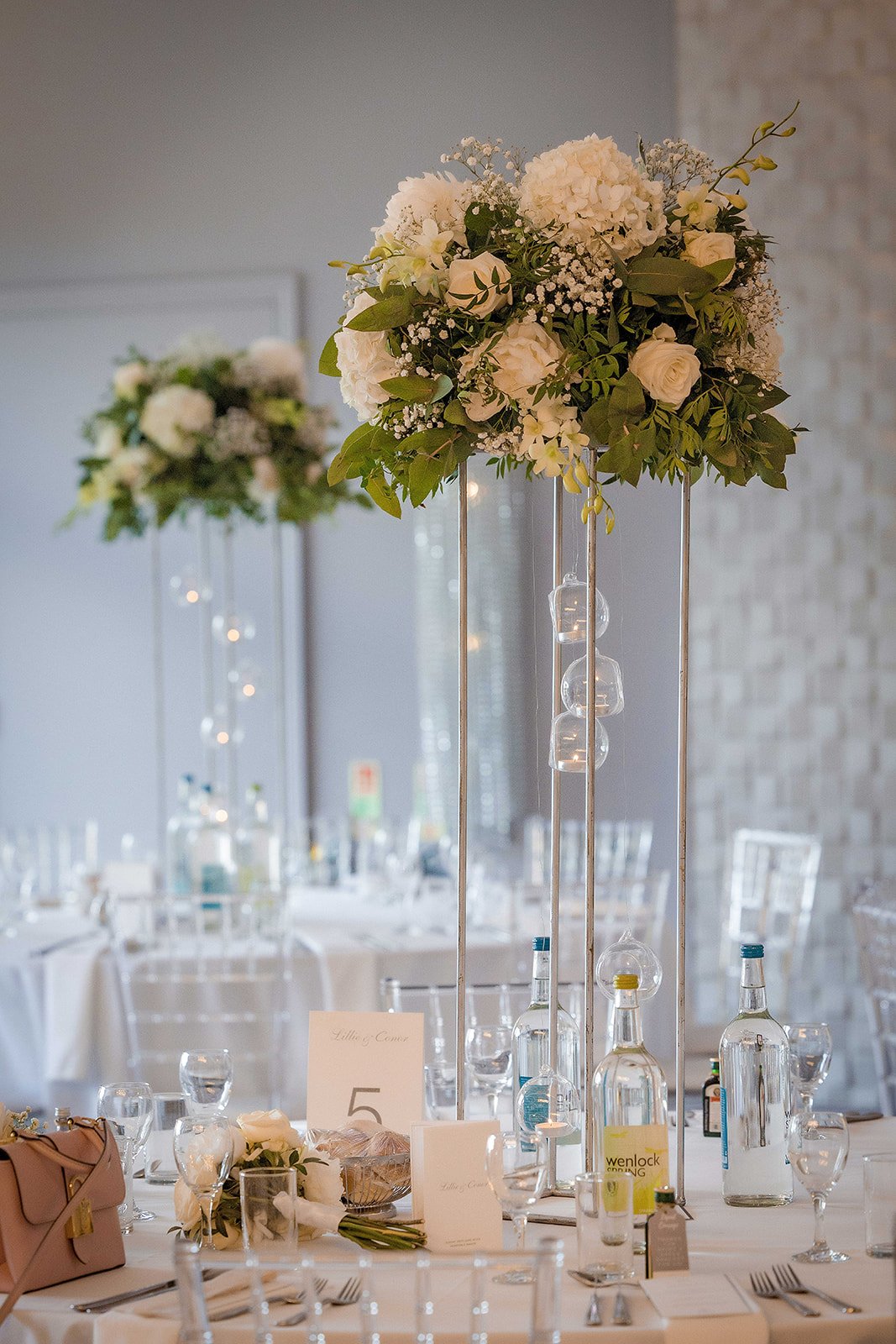 Elegant wedding reception table with tall floral centerpiece, glass vases, white roses, hydrangeas, and greenery, set with glassware, bottles, and table decor.