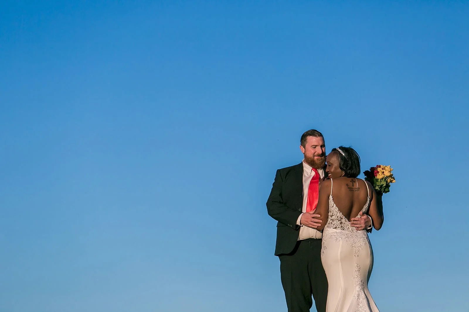 mixed race newlyweds couple embrace with clear blue sky background
