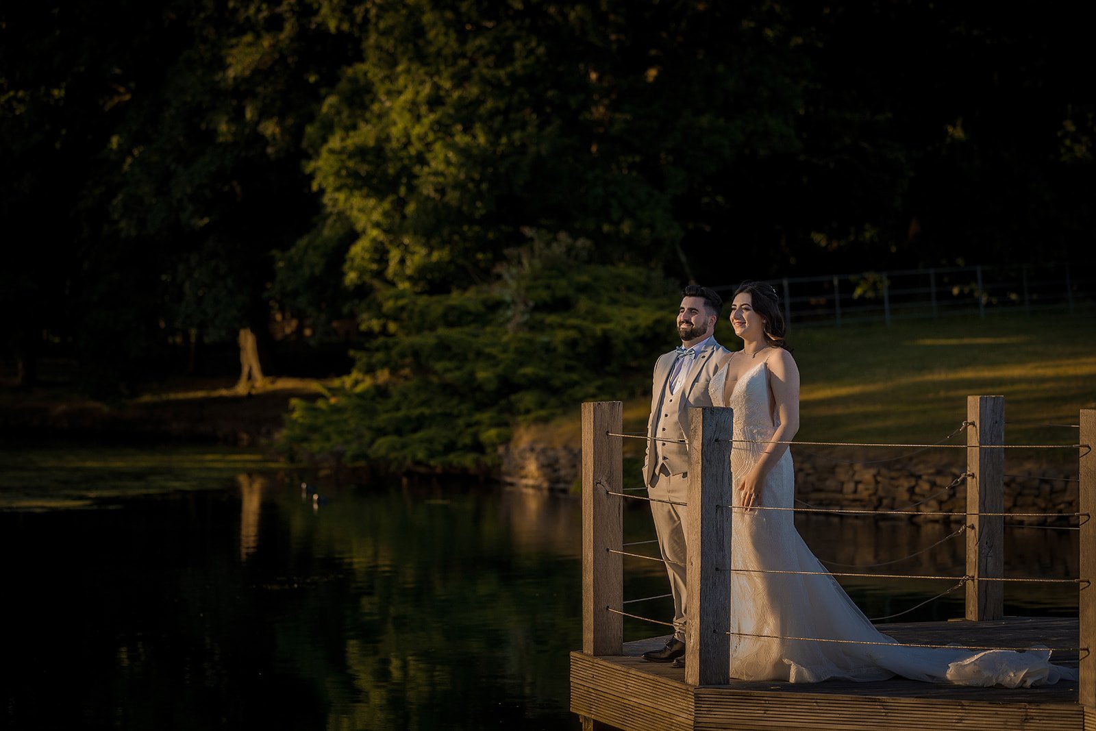 A newlywed couple at Merrydale Manor stands on a wooden dock by a lake at sunset. The groom is wearing a light gray suit with a bow tie, and the bride is in a white wedding gown. 