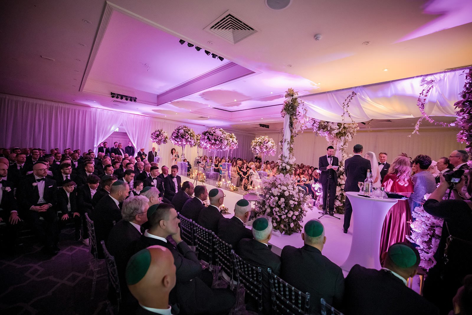Guests watching ceremony under purple uplighting and chandelier, The Mere reception suite
