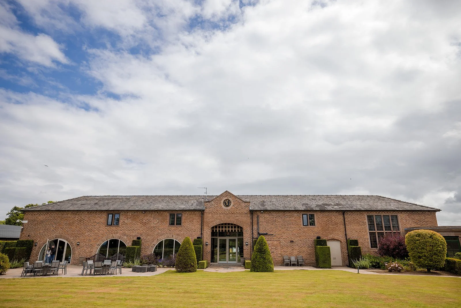 A large brick building with arched windows and a glass door, surrounded by well-manicured lawns and shrubs, with outdoor seating in front and a cloudy sky overhead.