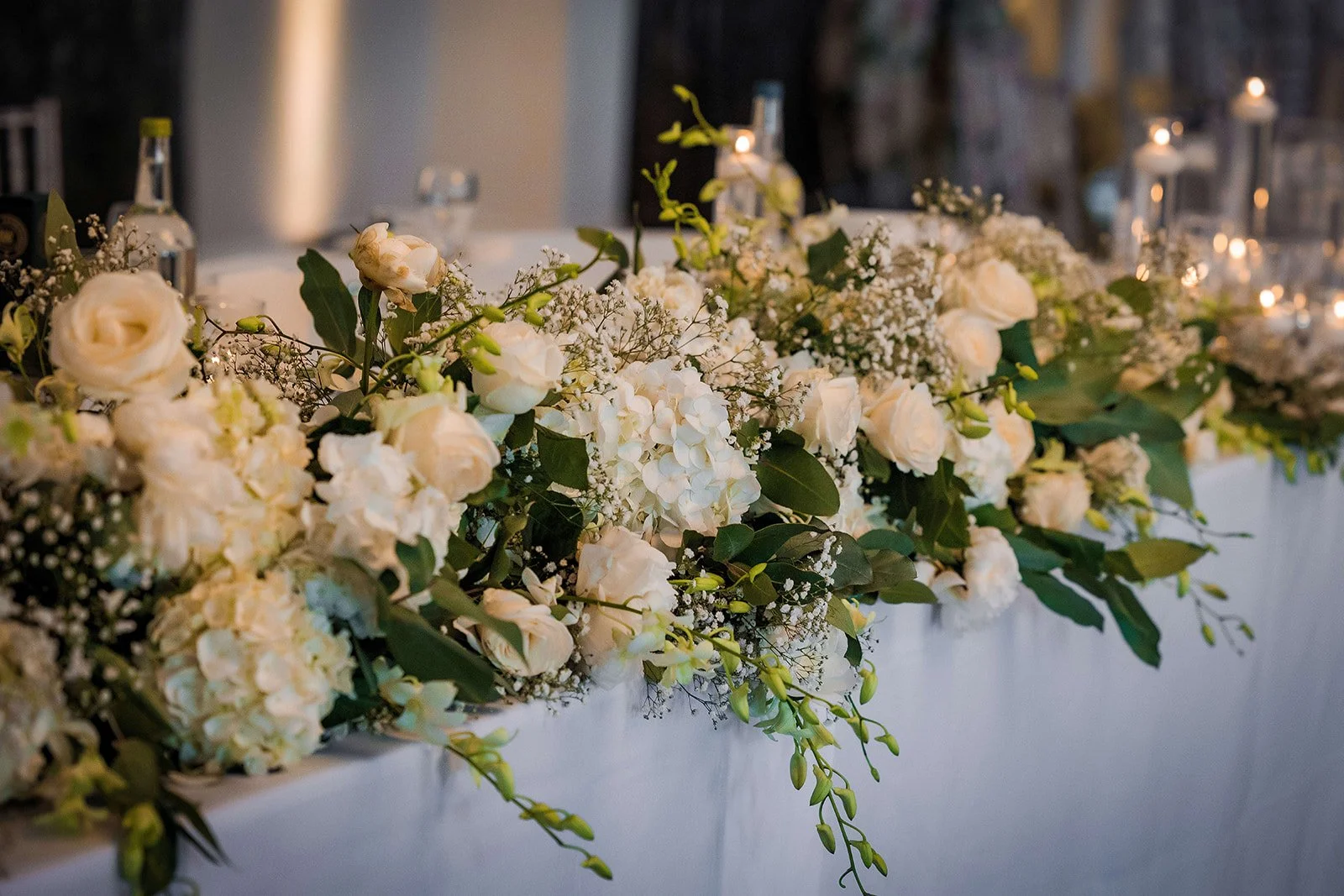 Close-up of a white floral garland with roses, hydrangeas, baby's breath, and greenery on a table, with candles in the background.