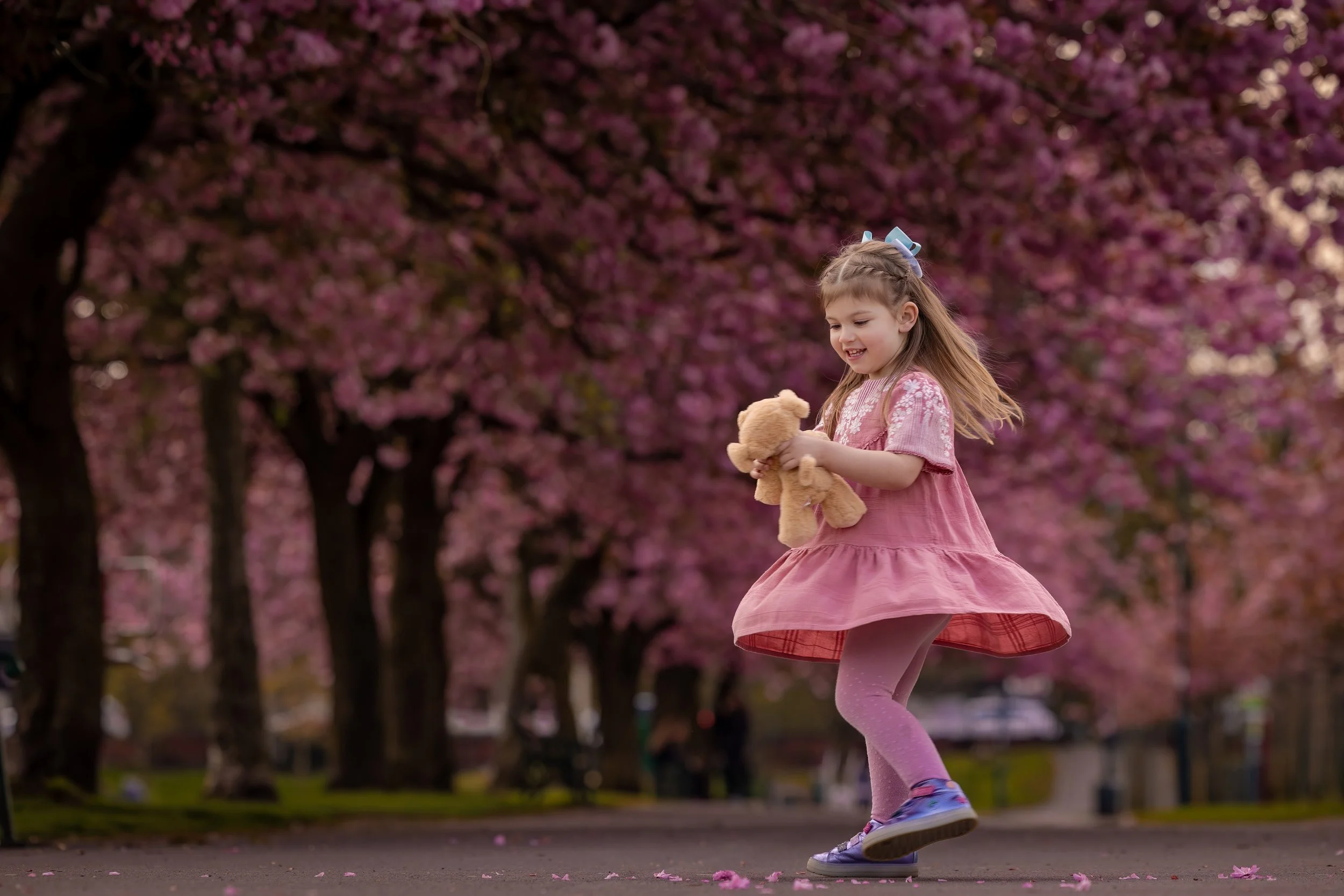A young girl in a pink dress and purple leggings roller-skates in a park, holding a teddy bear, with pink blossoms in the background.