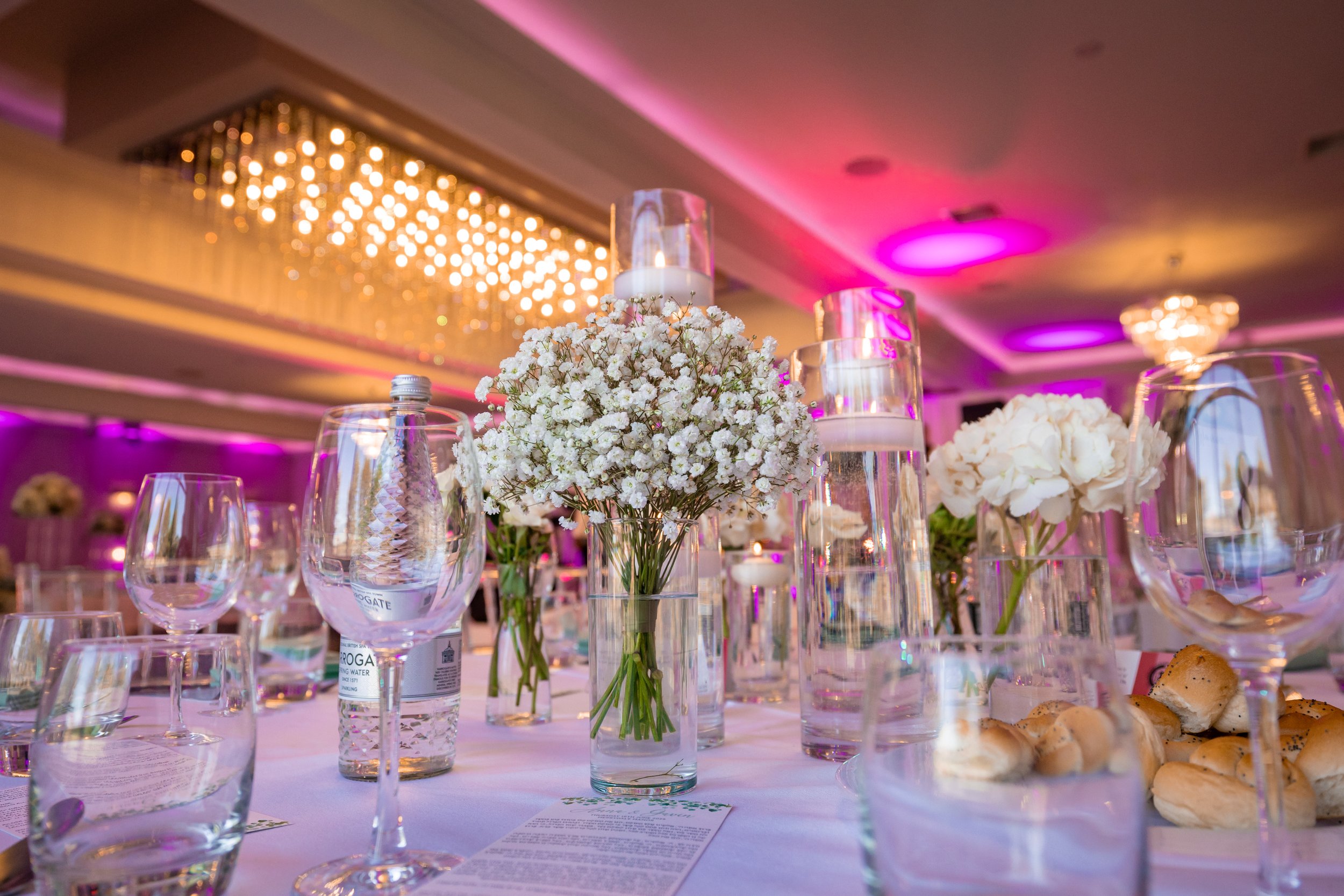 A decorated event table with multiple glasses, a floral arrangement of white flowers, and candles in glass holders, under pink and purple lighting.