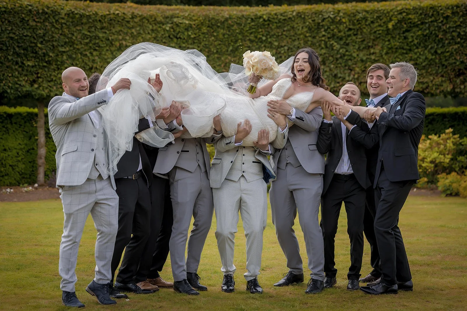 A group of groomsmen at Merrydale Manor lifting bride in a wedding dress holding a bouquet of white roses
