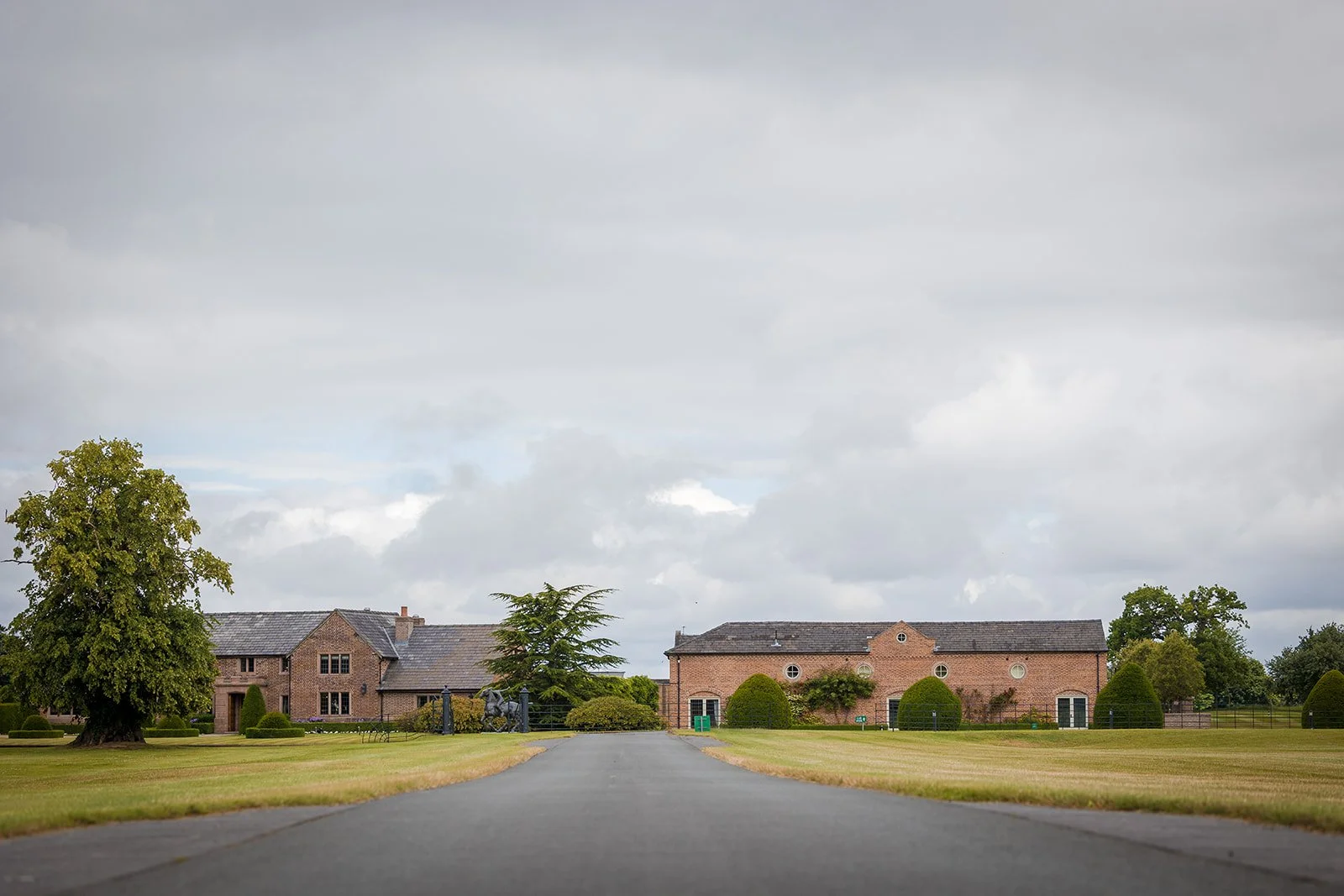 A long driveway leading to a large brick house with neatly trimmed bushes and trees on a cloudy day.