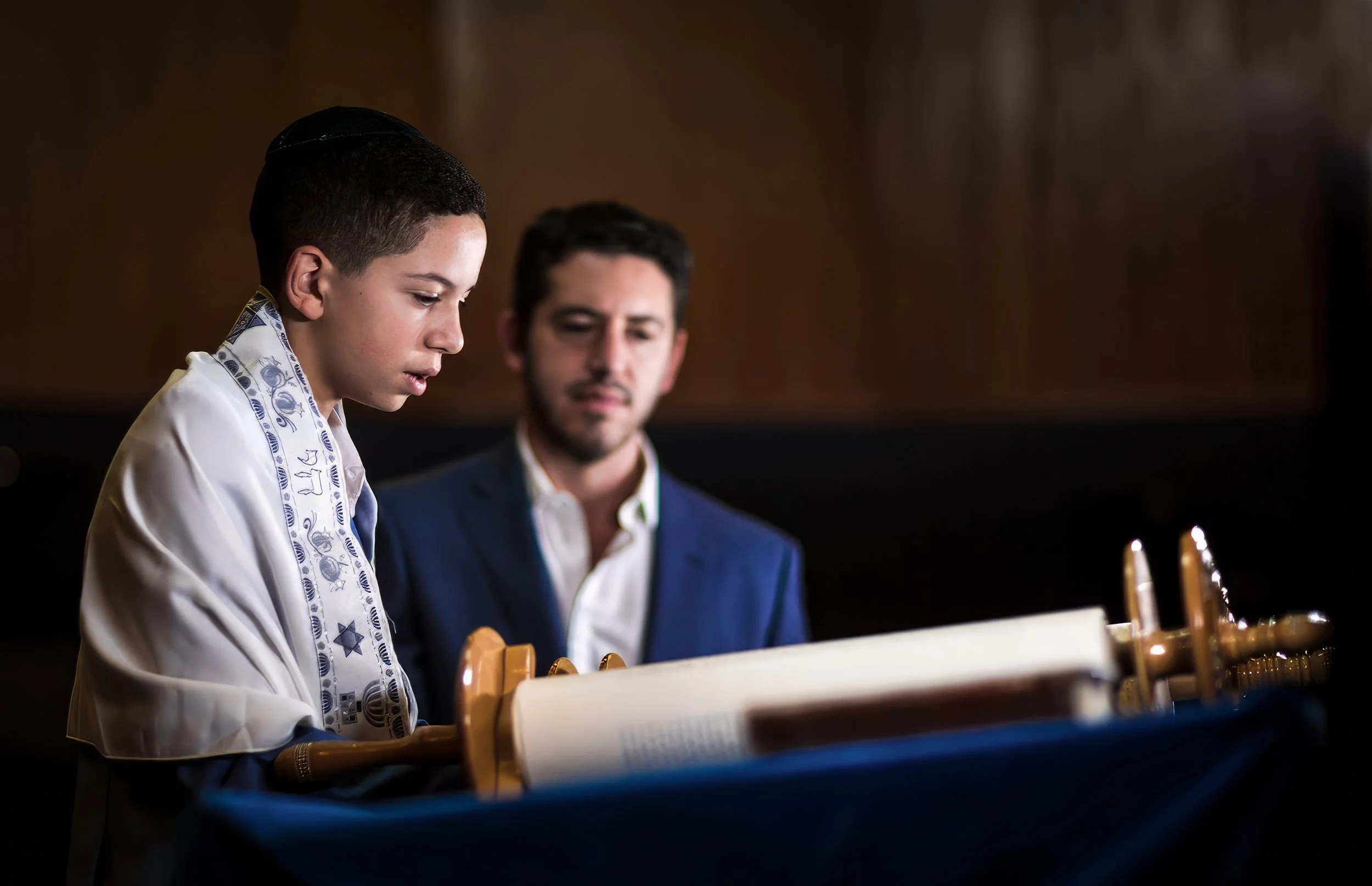 barmitzvah boy with tallit on reads from torah in synagogue watched by proud dad in Manchester