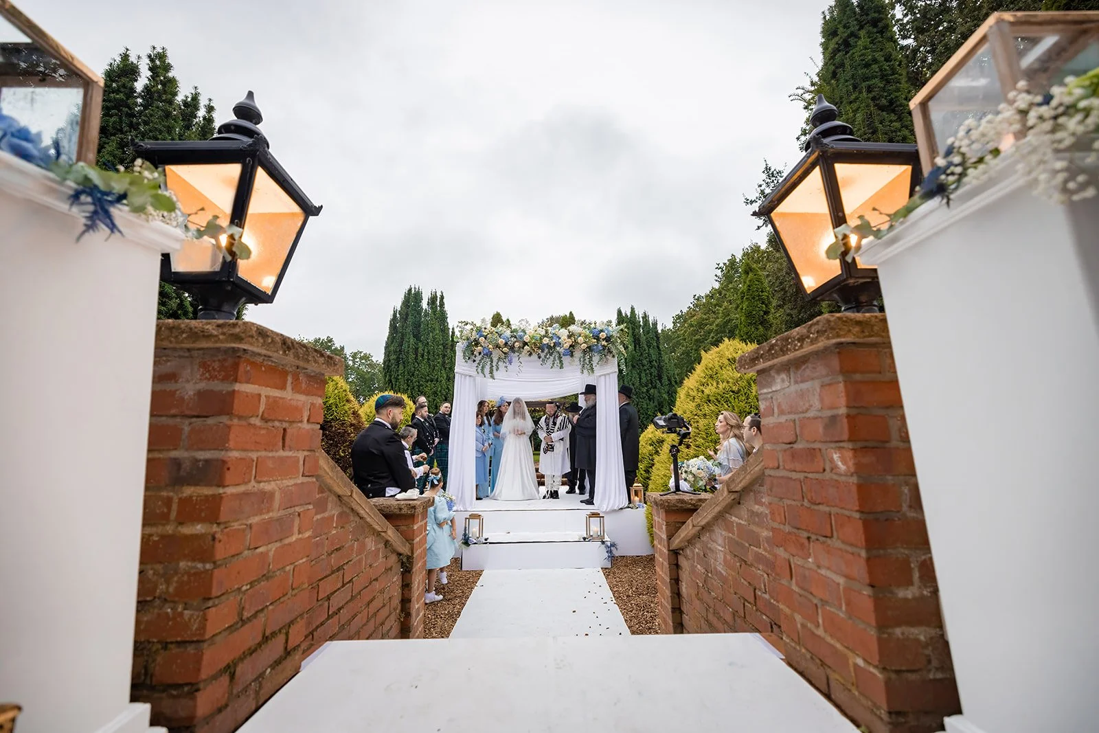 View down the white aisle between Nunsmere Hall's lantern-flanked entrance pillars, couple at altar
