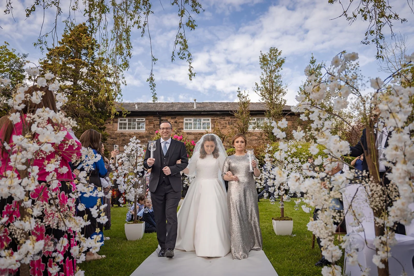 bride and parents make entrance down aisle to outdoor wedding ceremony at last drop village in bolton