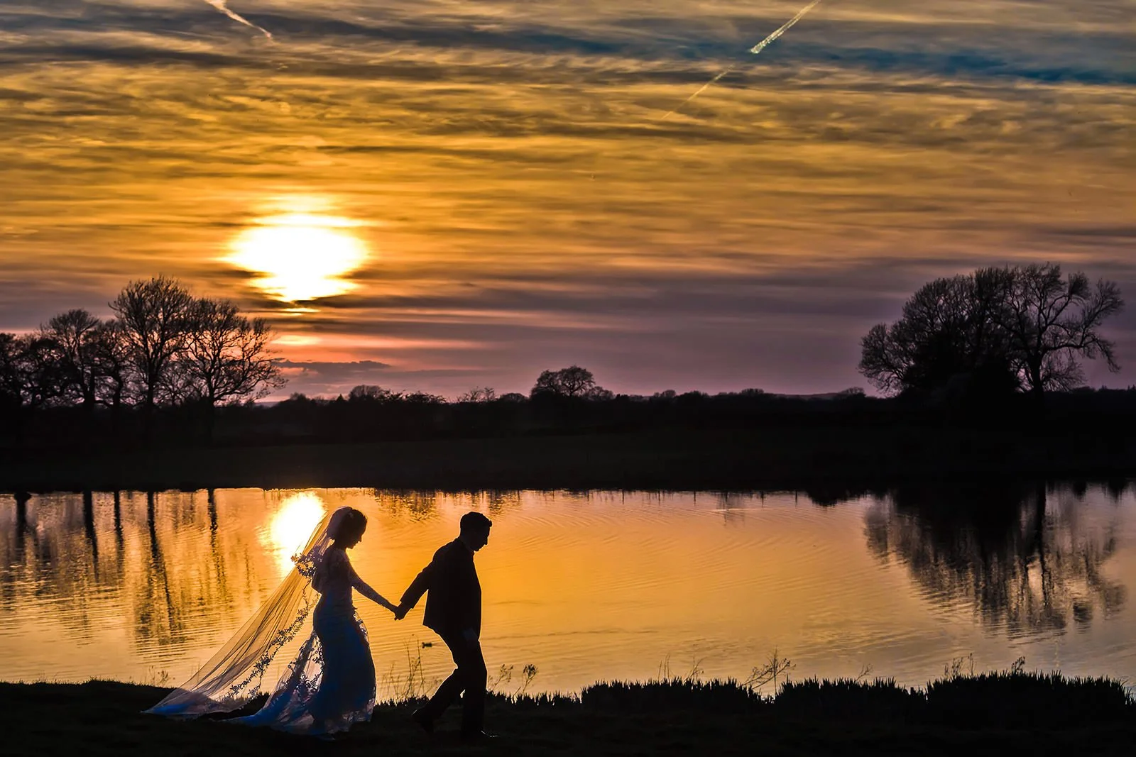 bride and groom silhouetted against lake with dramatic sunset at Styal Lodge in Manchester