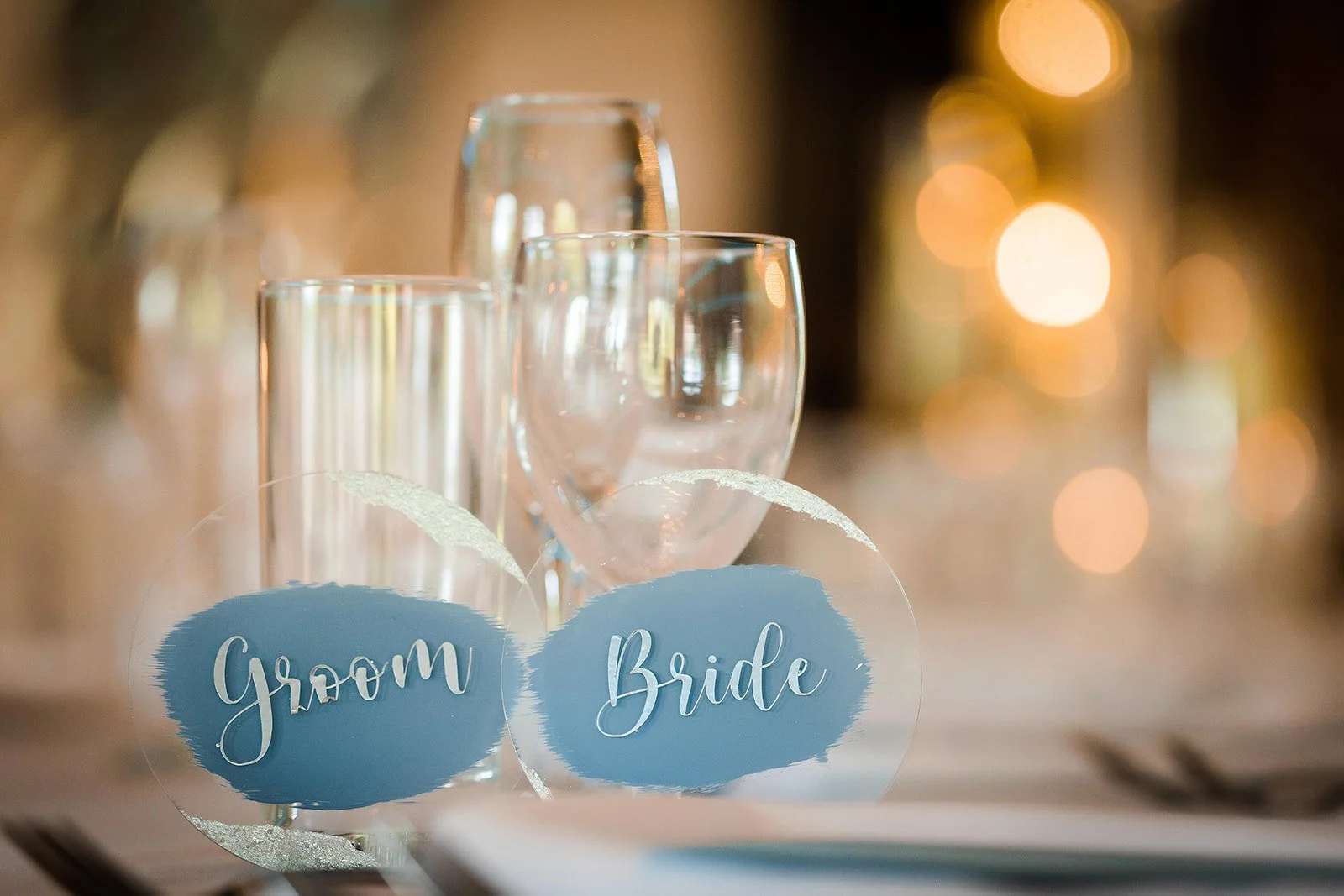 Glassware including a wine glass, a champagne flute, and a water glass on a table with blush pink and gold accents and a blurred background of round warm lights. Tags or labels reading 'Groom' and 'Bride' are also on the table.