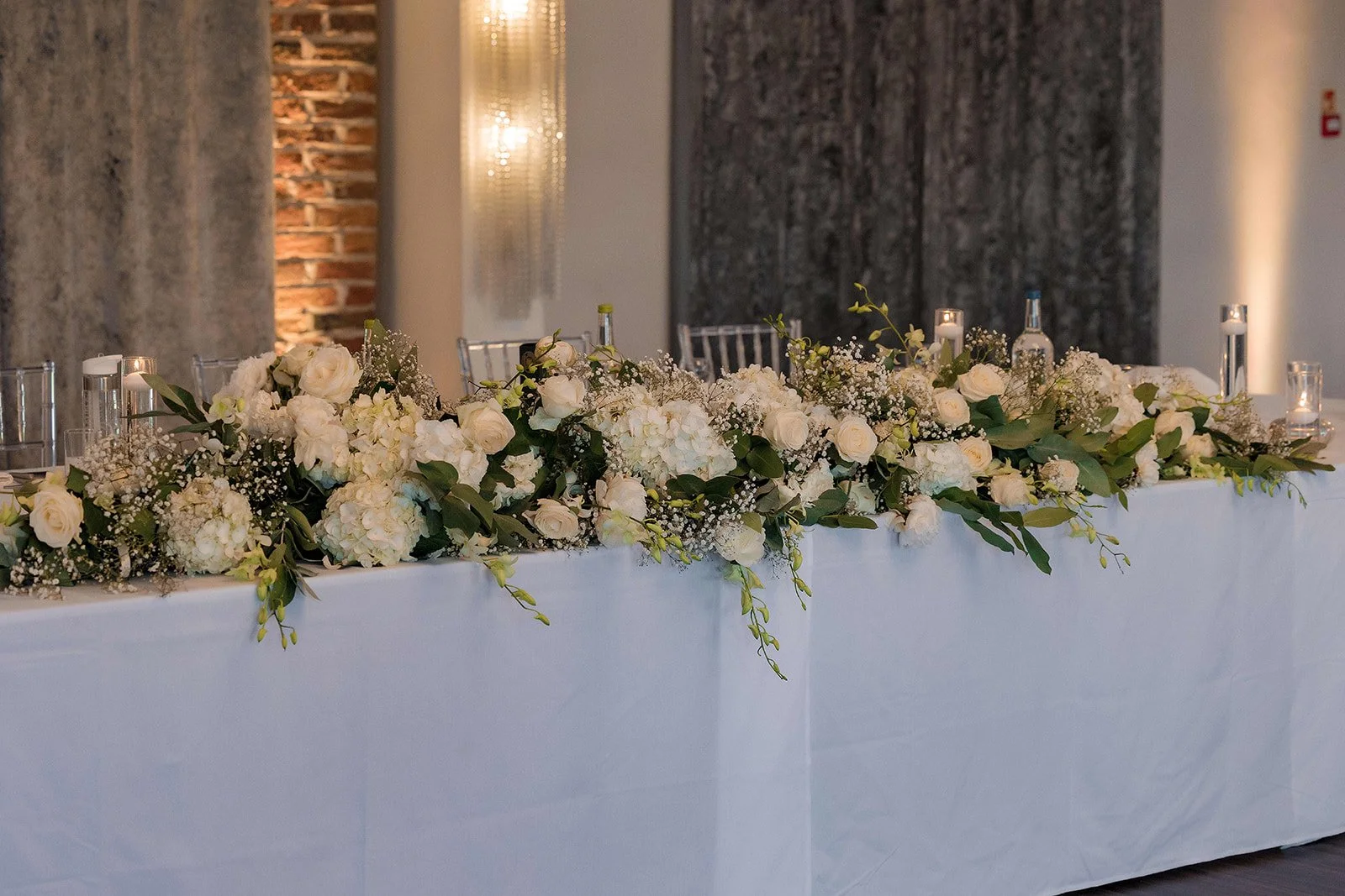 A long table decorated with white flowers including roses and hydrangeas, greenery, candles in glass holders, and silver chairs in the background at an indoor event space.