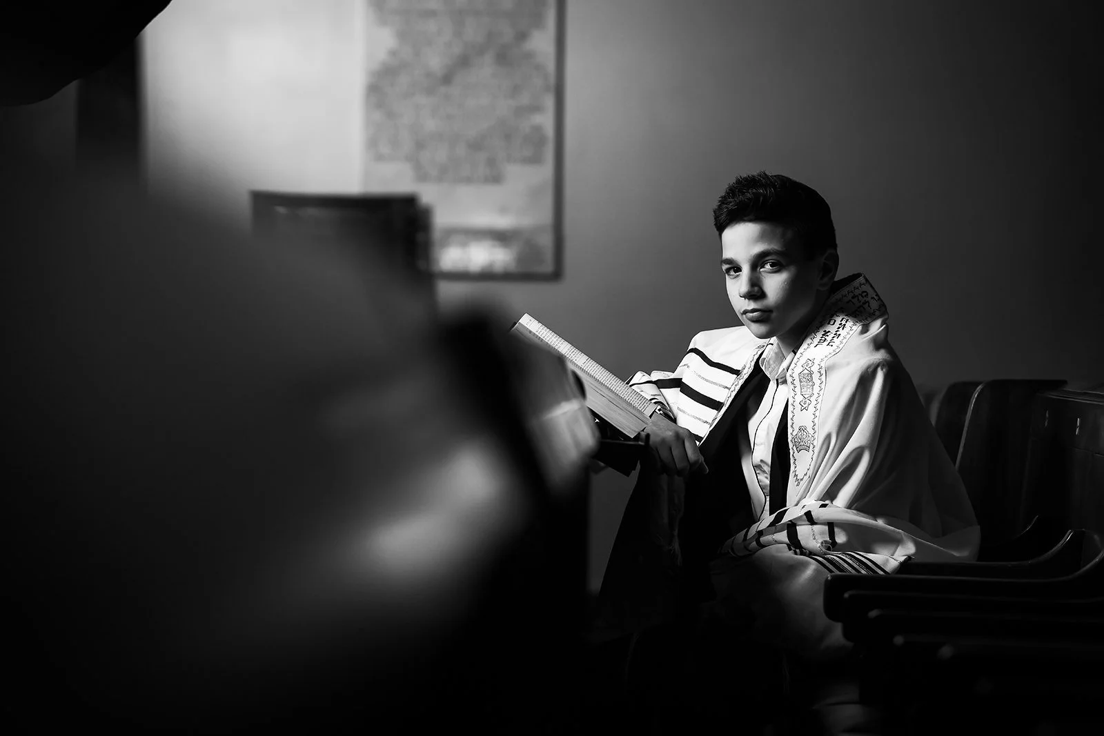 black and white posed portrait of barmitzvah boy sitting down in synagogue with prayer book
