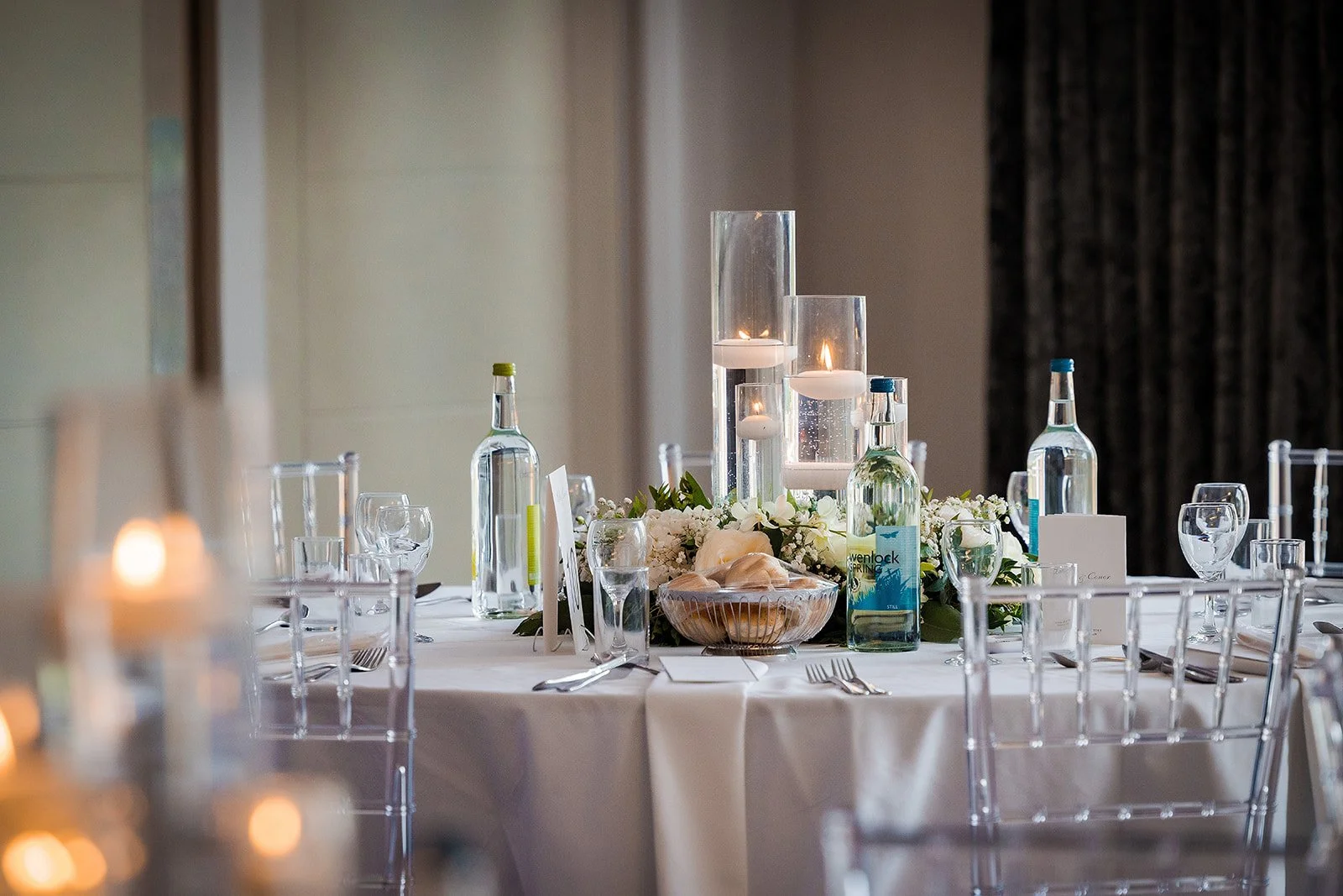 A round banquet table decorated with white tablecloths, clear chairs, and a floral centerpiece with candles, wine bottles, and glassware, set for an event or celebration.