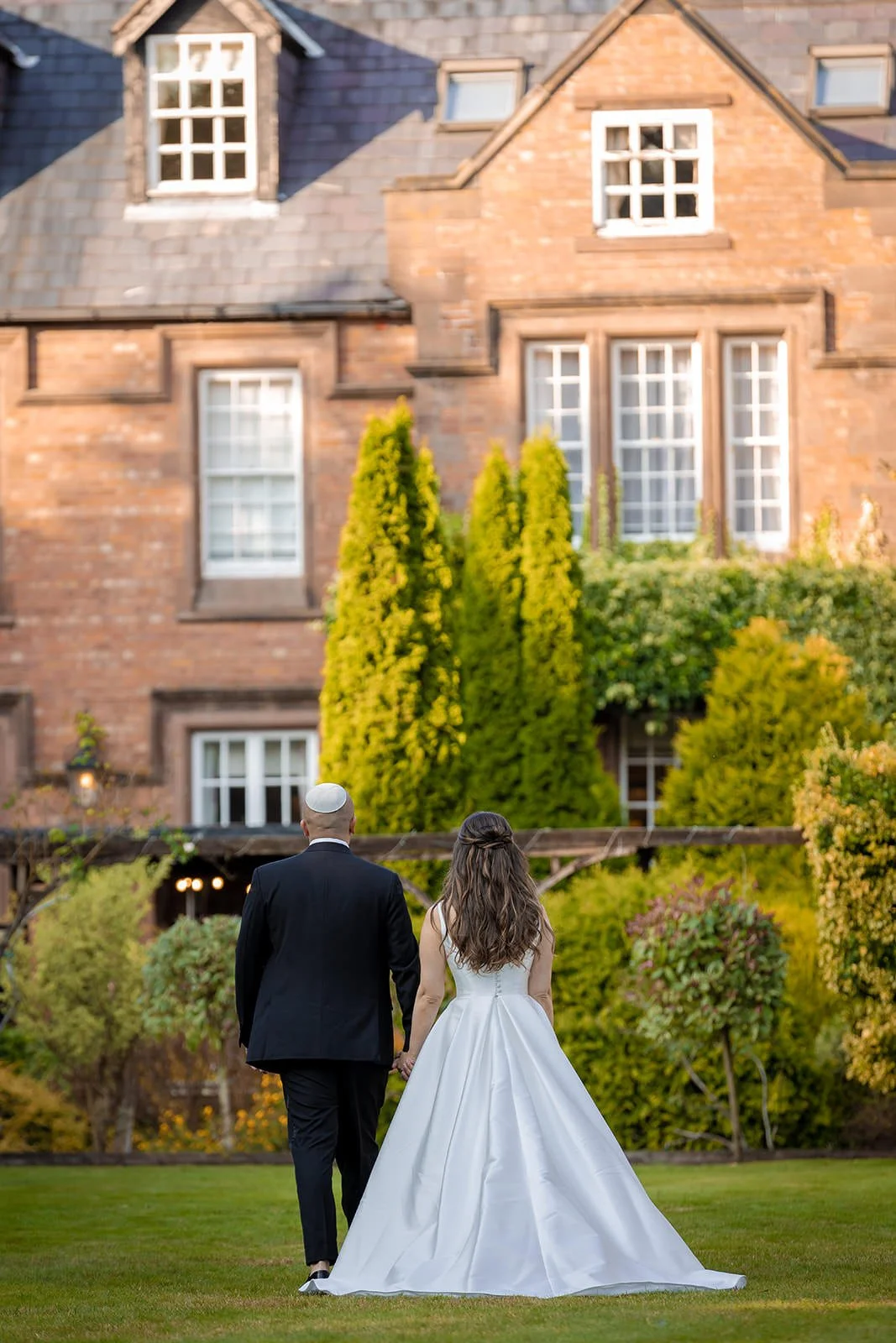 Bride and groom walking toward Nunsmere Hall manor house, autumn gardens
