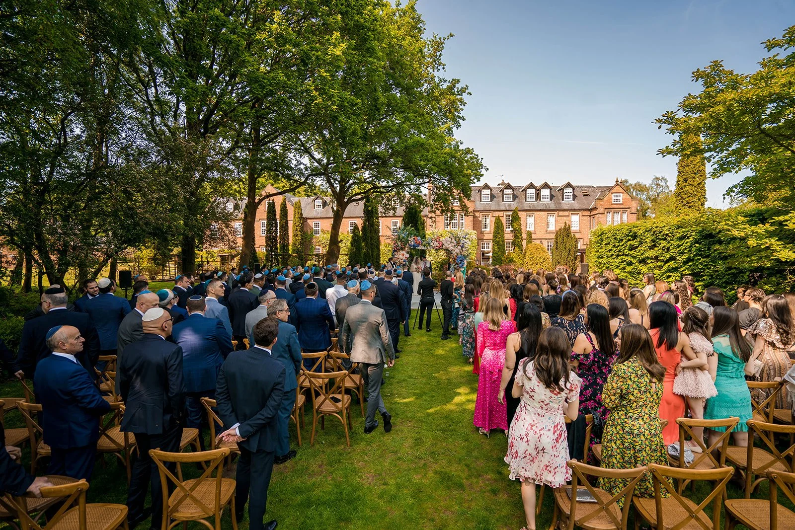 Wedding guests watching outdoor ceremony on the lawn at Nunsmere Hall, manor house behind


