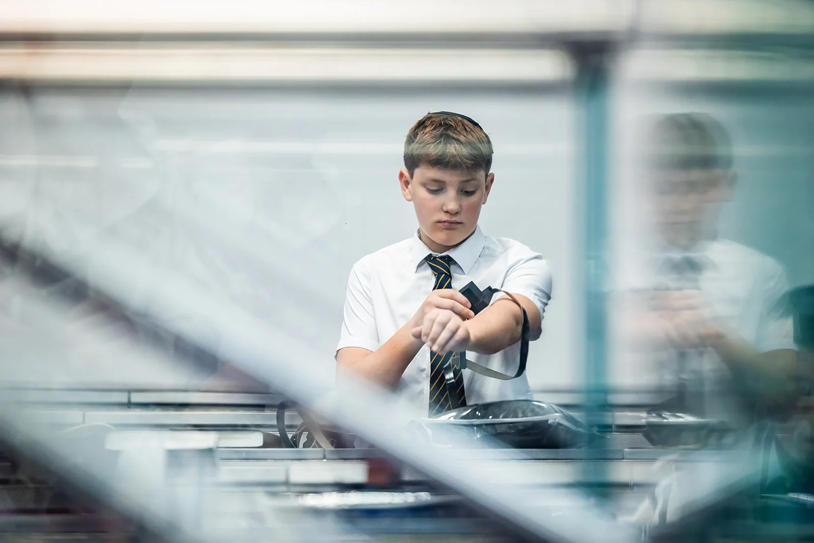 barmitzvah boy puts on tefillin in synagogue framed by glass