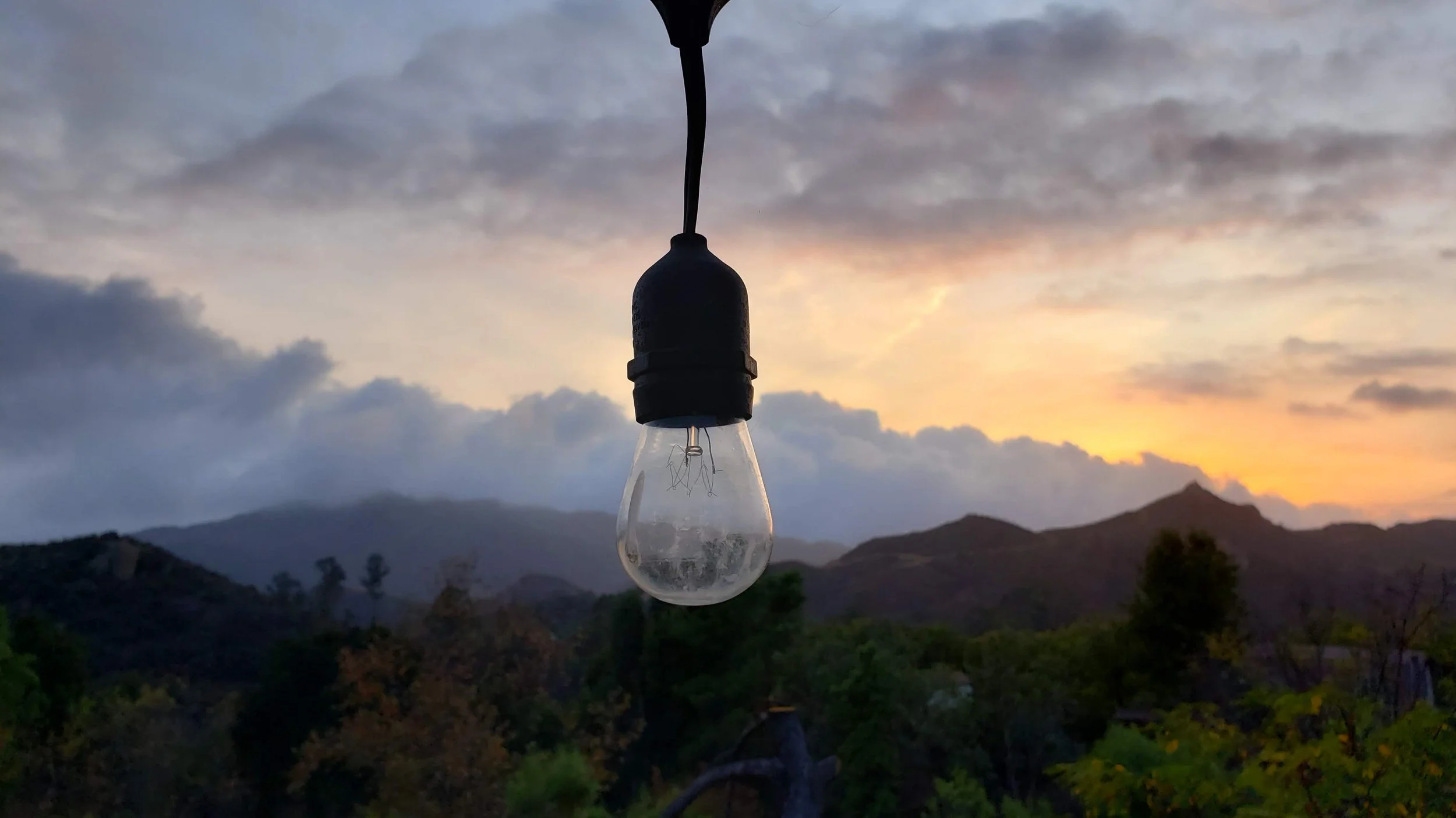 A clear light bulb hanging from a black socket against a sunset sky with mountains in the background.