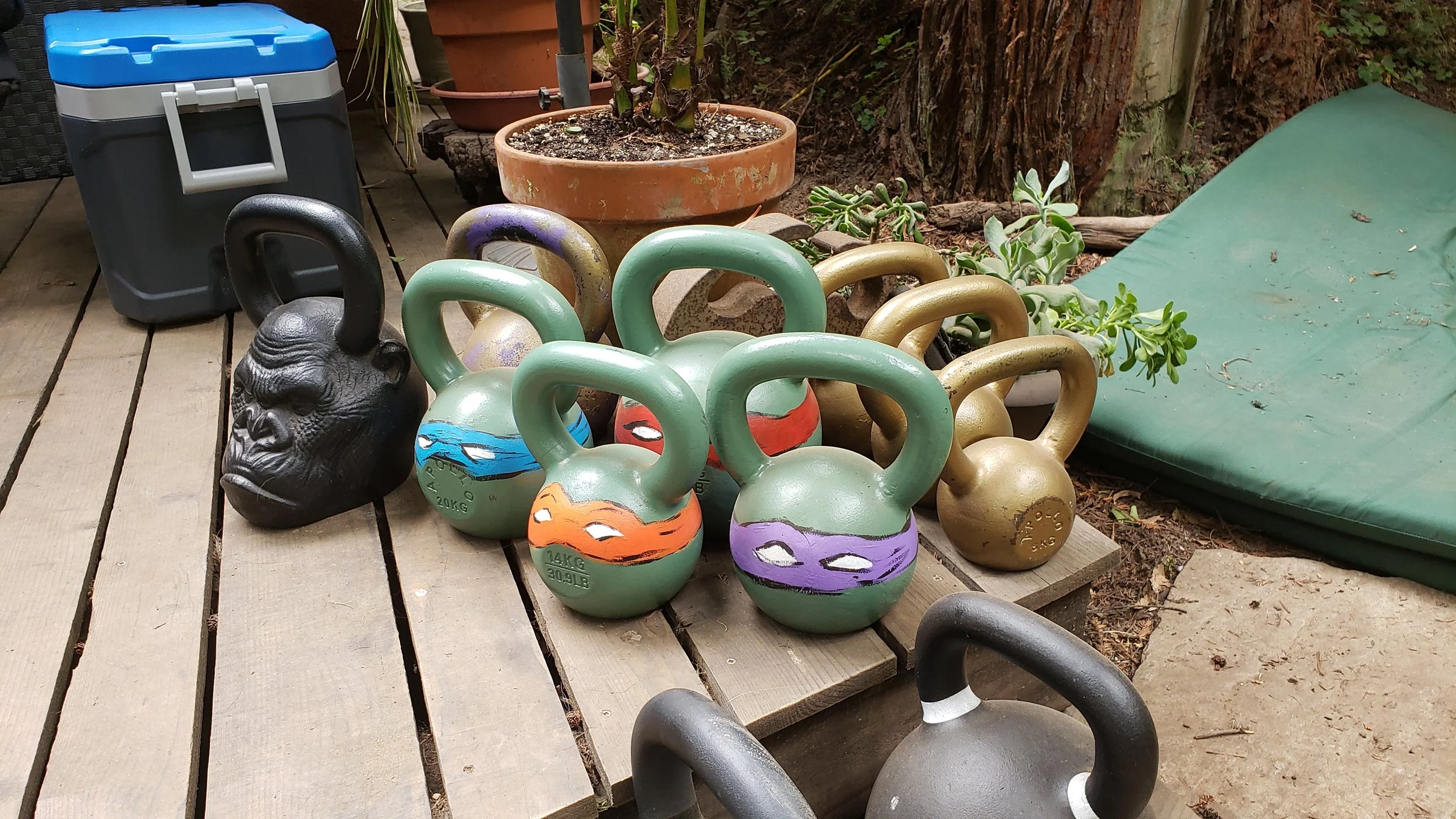 Outdoor scene with colorful kettlebells, a decorative black gorilla head, a cooler, potted plants, and a green outdoor cushion on a wooden deck.