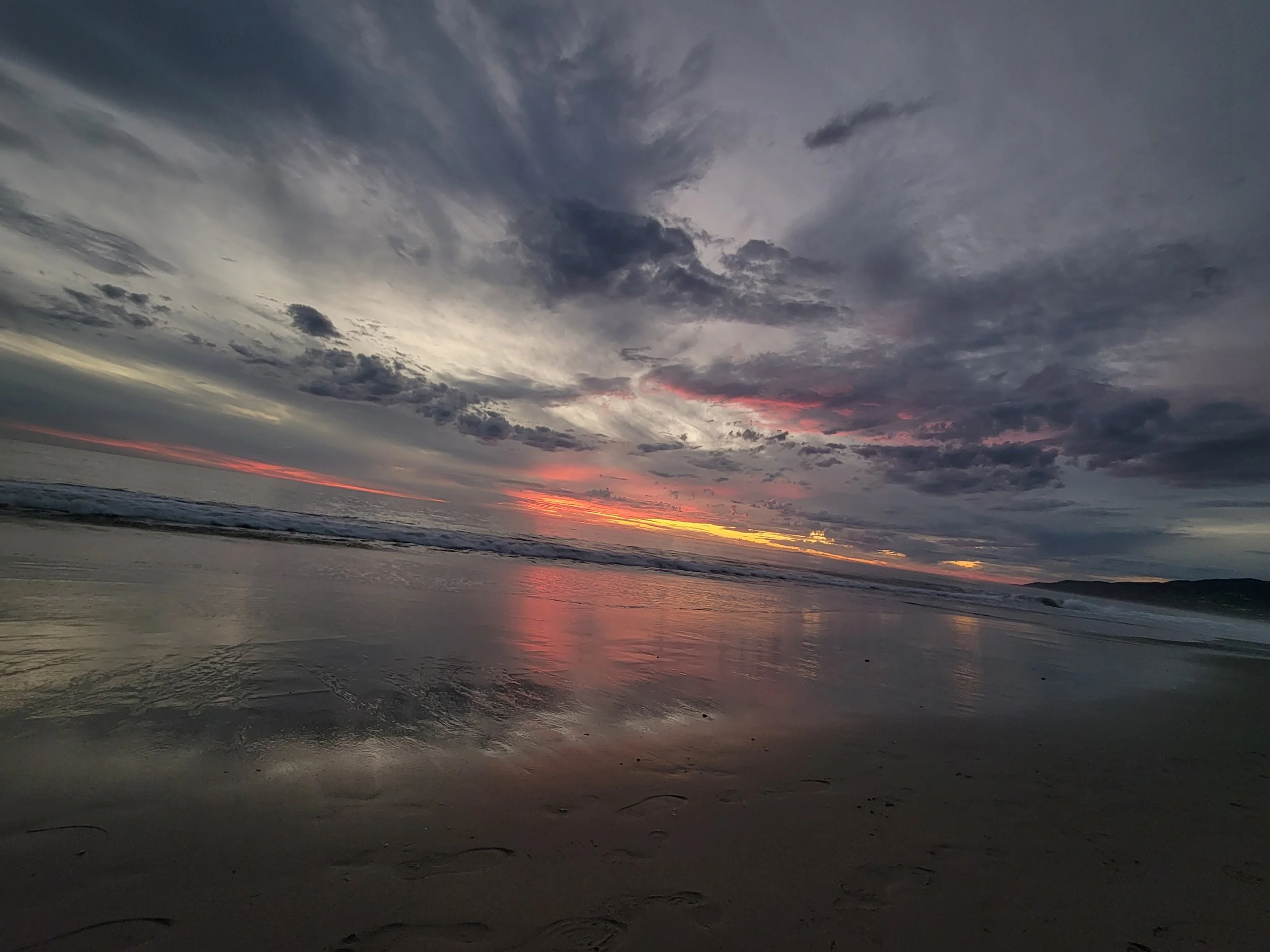 A beach at sunset with a cloudy sky, colorful horizon, and wet sand reflecting the sky.