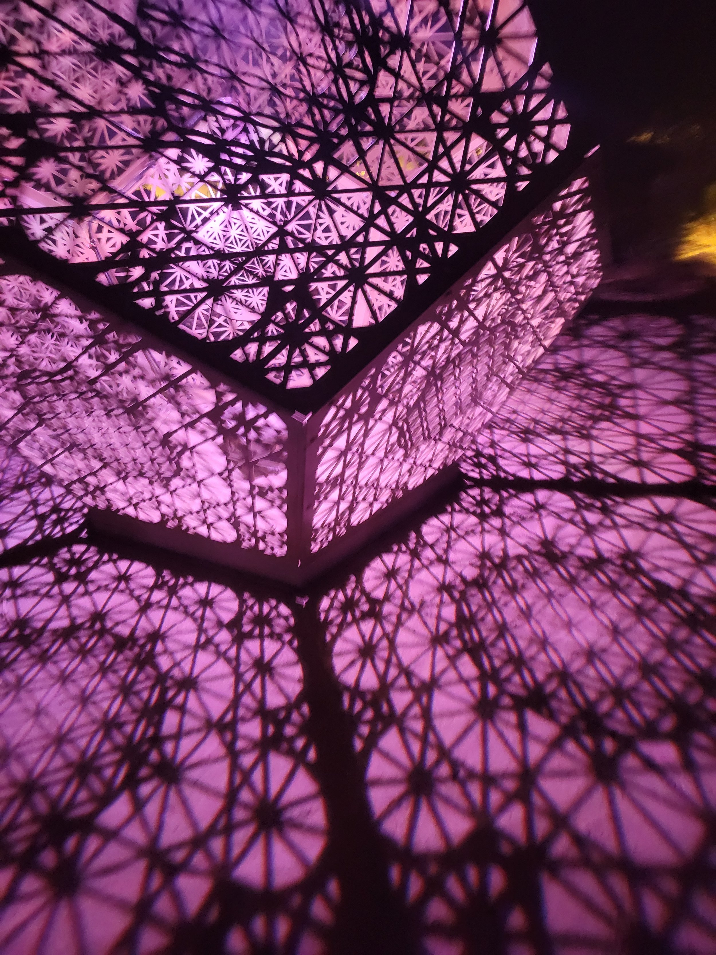 Close-up view of a structure with a geometric, lattice-like pattern illuminated in purple light, creating intricate shadows.