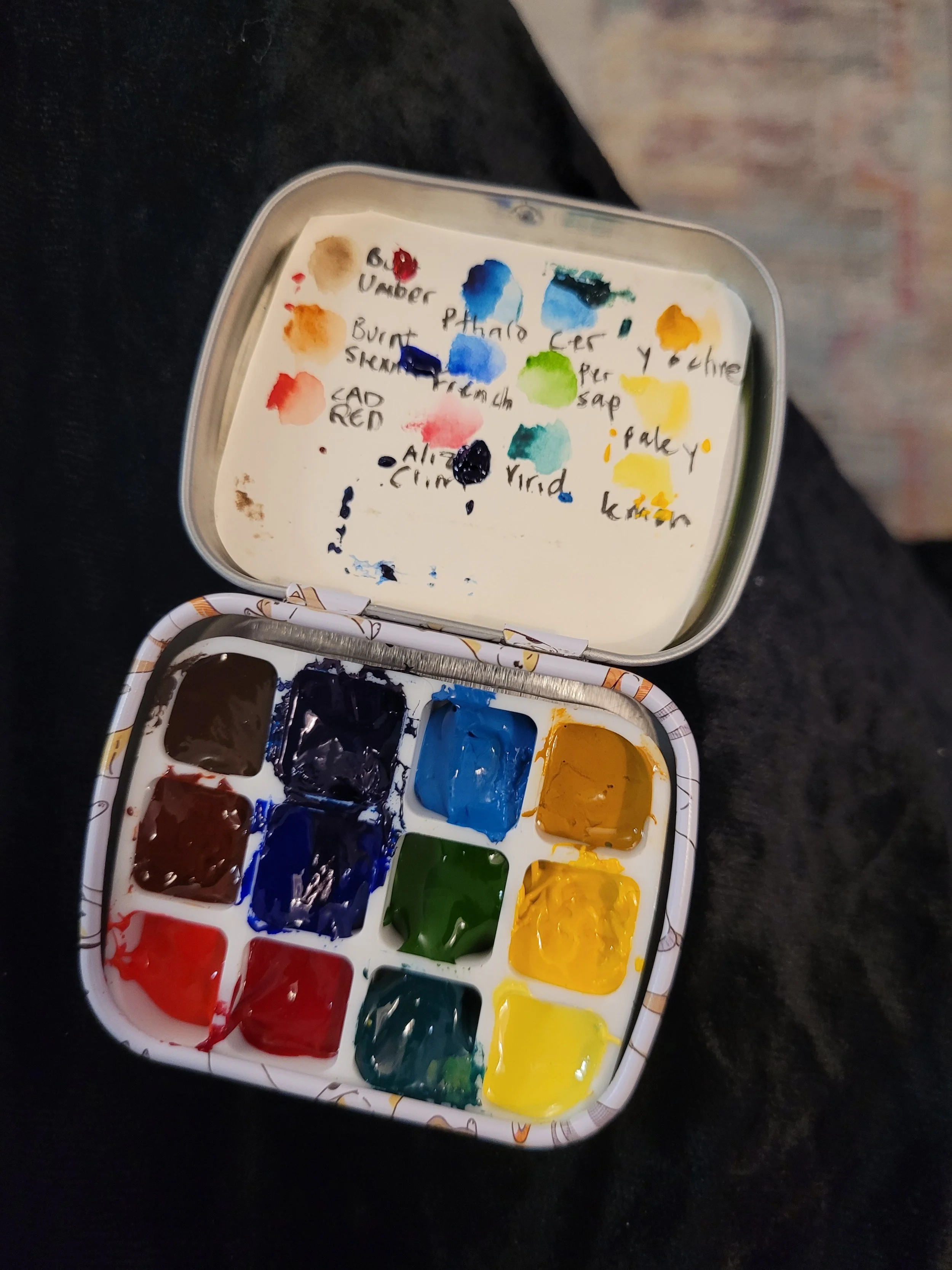A watercolor paint palette with red, blue, green, yellow, and brown colors, and a sheet of paper with handwritten color names and smeared paint swatches.