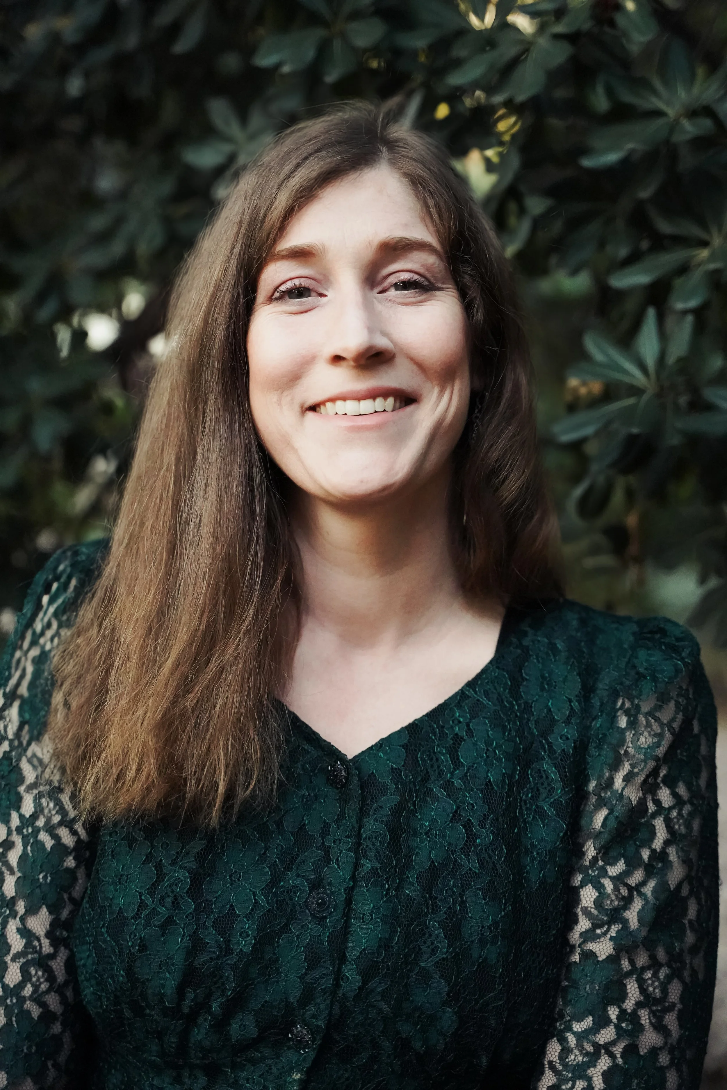 A woman with long brown hair and light skin, smiling, wearing a dark green lace dress, standing outdoors with green foliage in the background.
