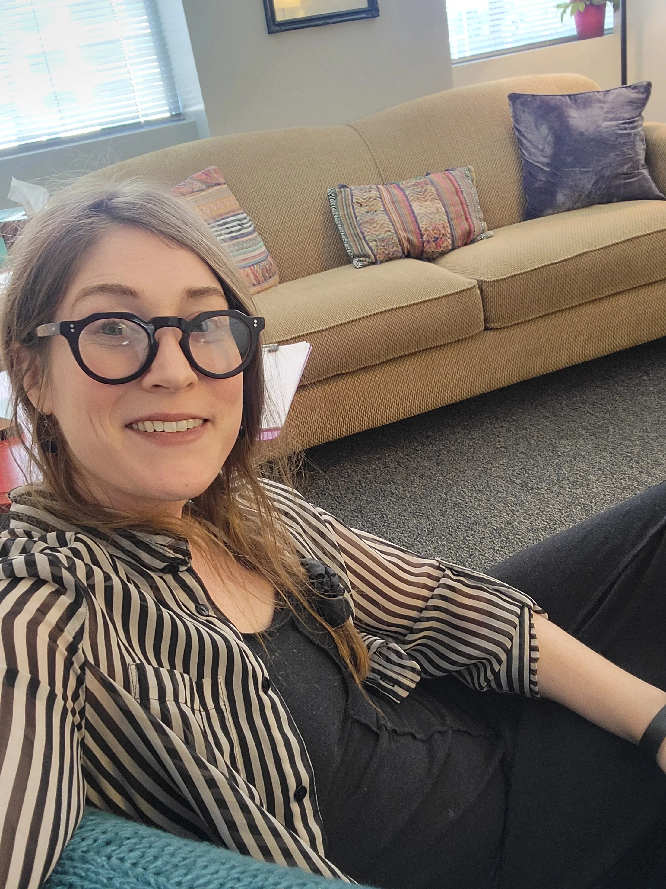 A woman with long blonde hair, wearing black-rimmed glasses and a black and white striped shirt, sitting on a black sofa in a room with beige walls, colorful cushions, and windows with blinds.