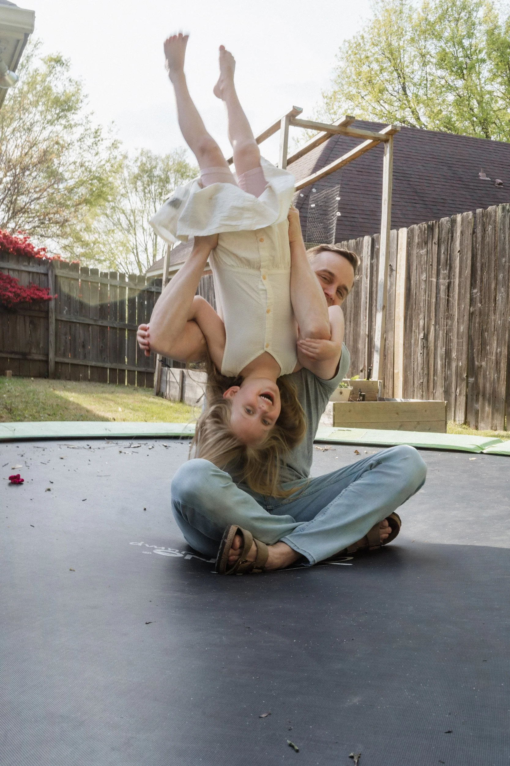 A man is sitting cross-legged on a trampoline, smiling, while holding a young girl upside down by her waist. The girl, with long hair and wearing a light-colored dress with pink shorts, is laughing and hanging upside down. The scene is set outdoors in a backyard with a wooden fence, trees, and a house with a roof in the background.