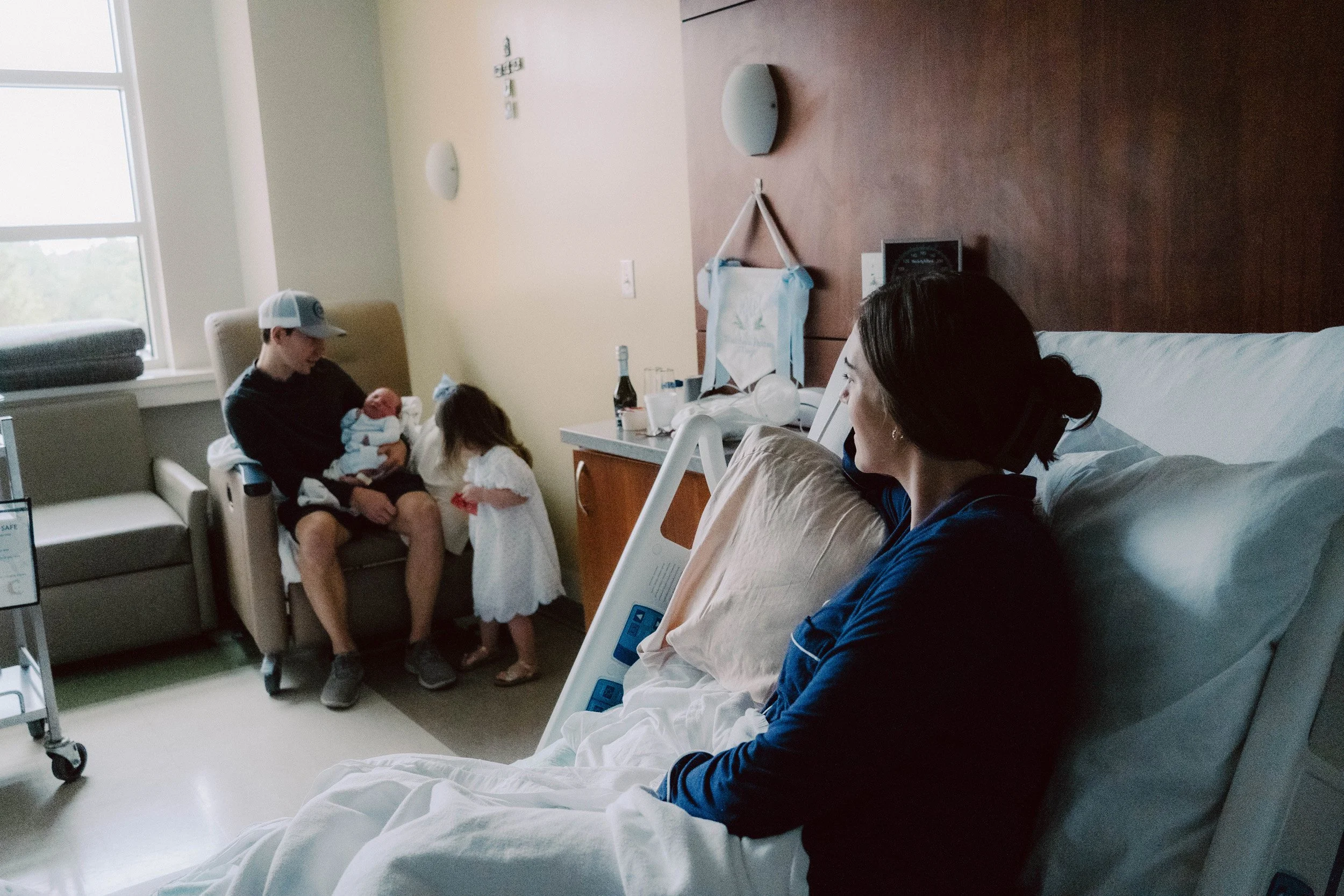 In a hospital room, a woman is sitting on a bed, watching a man hold a newborn baby. Two young girls are standing near the man, looking at the baby. The scene is bright with natural light from the window, and medical equipment is visible in the room.