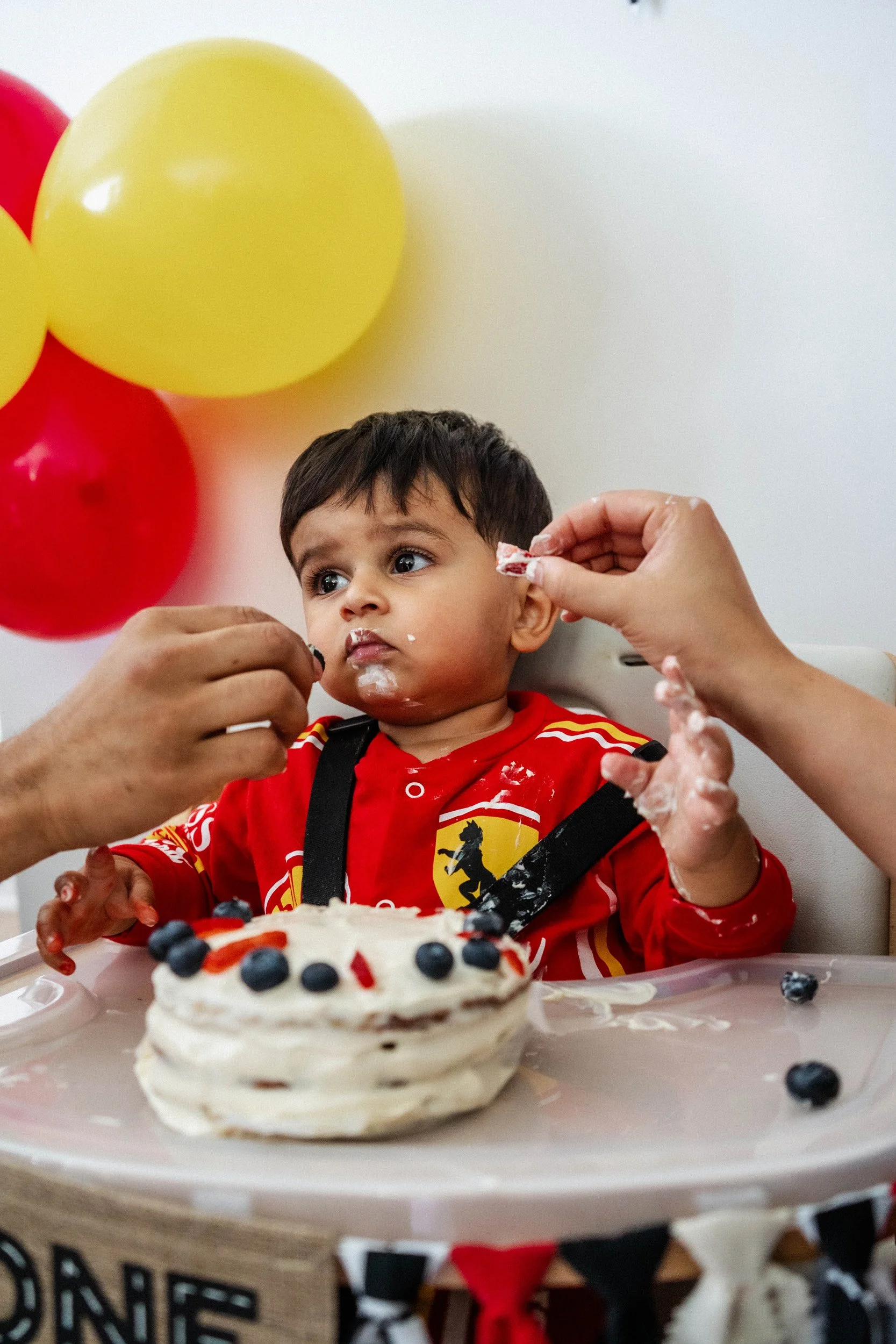 Young boy in a red superhero-themed shirt celebrating his birthday at a party with balloons and cake, with an adult hand feeding him a piece of cake.