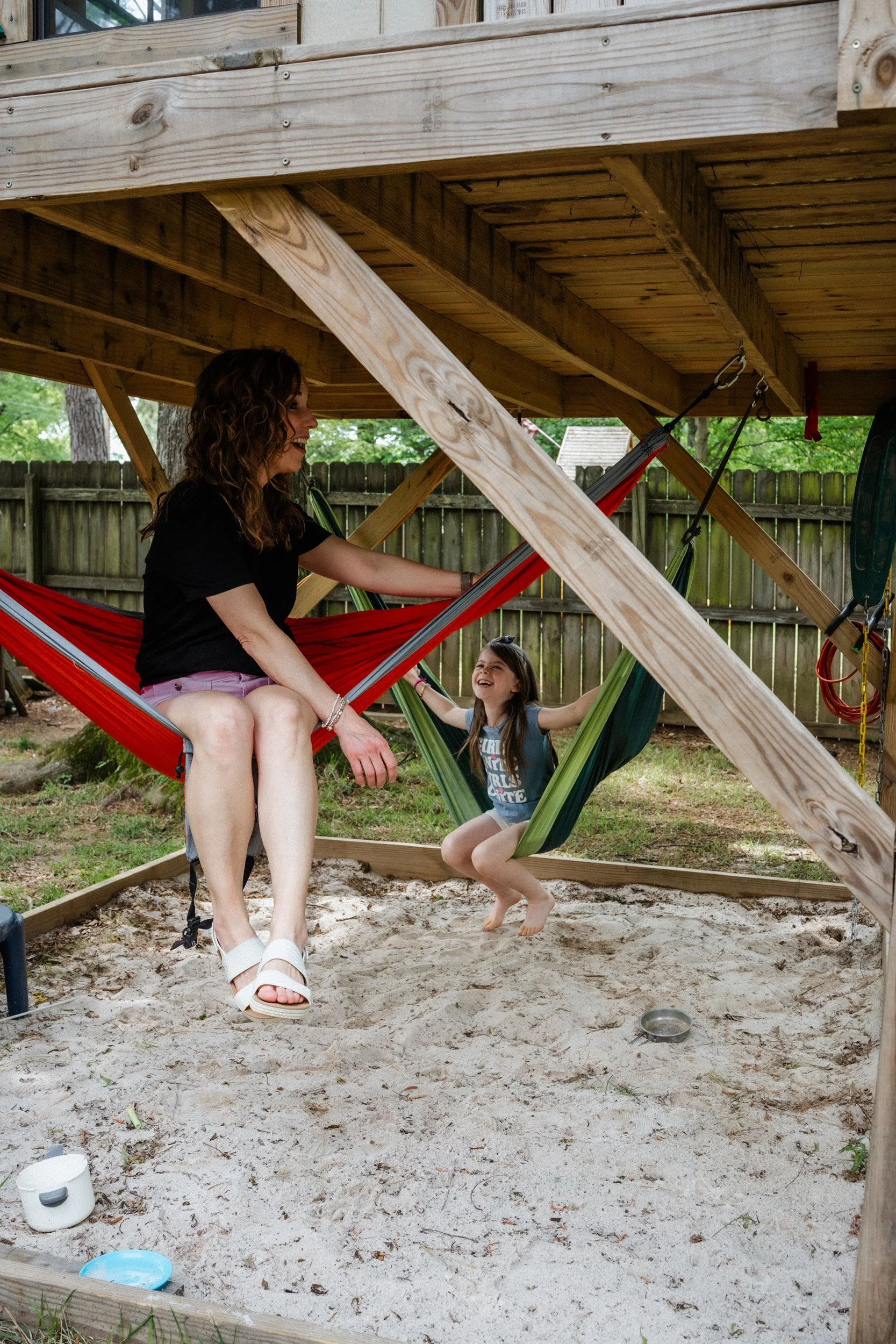 A woman and a young girl sitting on a hammock under a wooden deck in a backyard. The woman is smiling, wearing a black shirt and shorts, and white sandals. The girl is also smiling, wearing a graphic t-shirt and shorts, with arms outstretched. The ground is sandy, with some scattered toys and a fence in the background.
