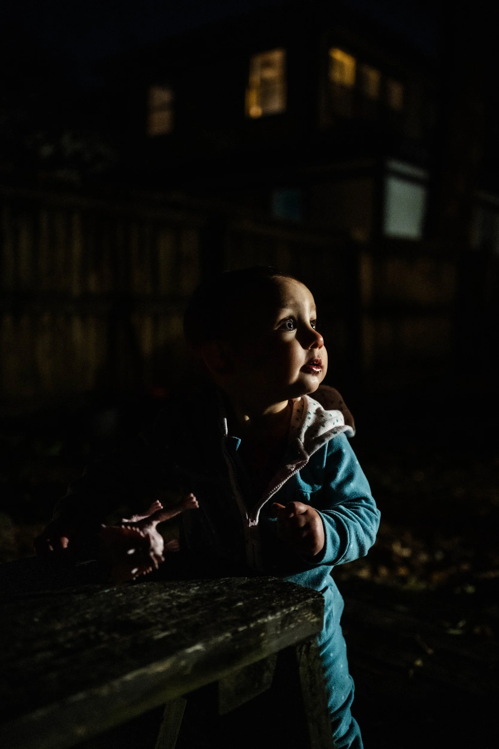 Young child with light skin and short hair, sitting outdoors at night, illuminated by a soft light, wearing a blue jacket with a hood and patterned shirt, with a wooden fence and house windows in the background.