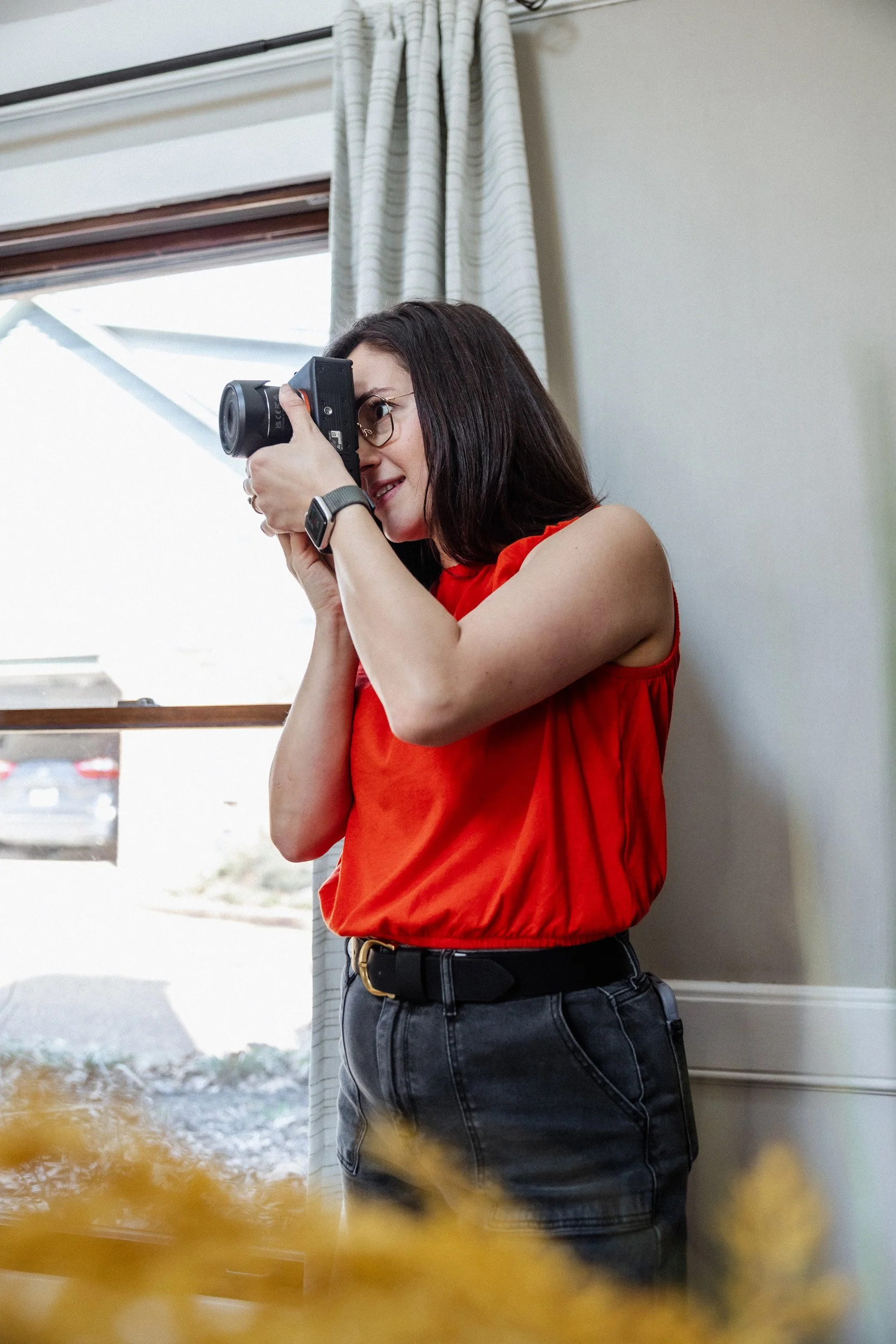 A woman with dark hair and glasses wearing a red sleeveless top and dark jeans is taking a photograph indoors, standing near a window with a white curtain.