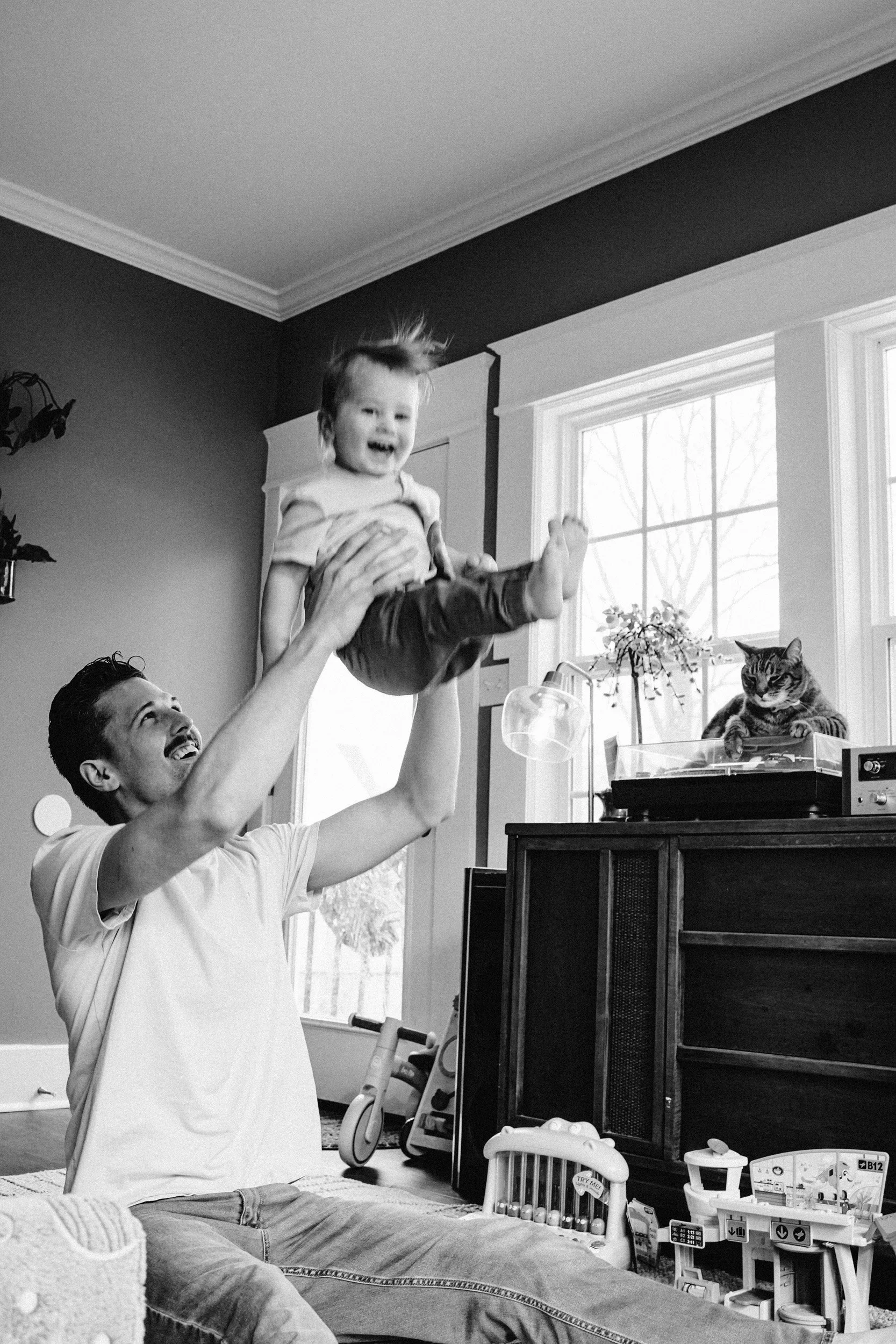 A man with a mustache sitting on the floor and holding a smiling young boy up in the air. The boy appears to be happy and having fun. The room has a window with sunlight coming in, and various toys and objects are scattered around.